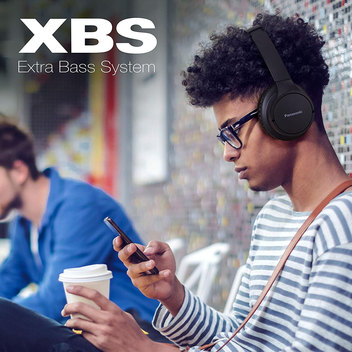 Młody mężczyzna ze słuchawkami i okularami używa telefonu. Osoba w tle.