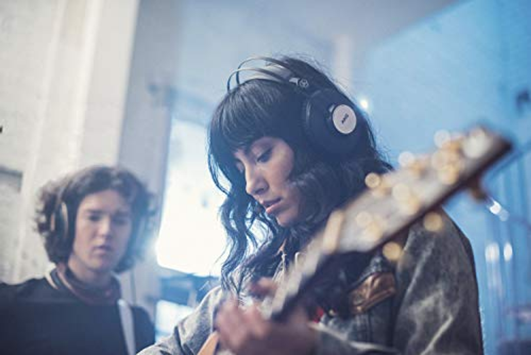 Donna che suona la chitarra con le cuffie, un'altra persona dietro.