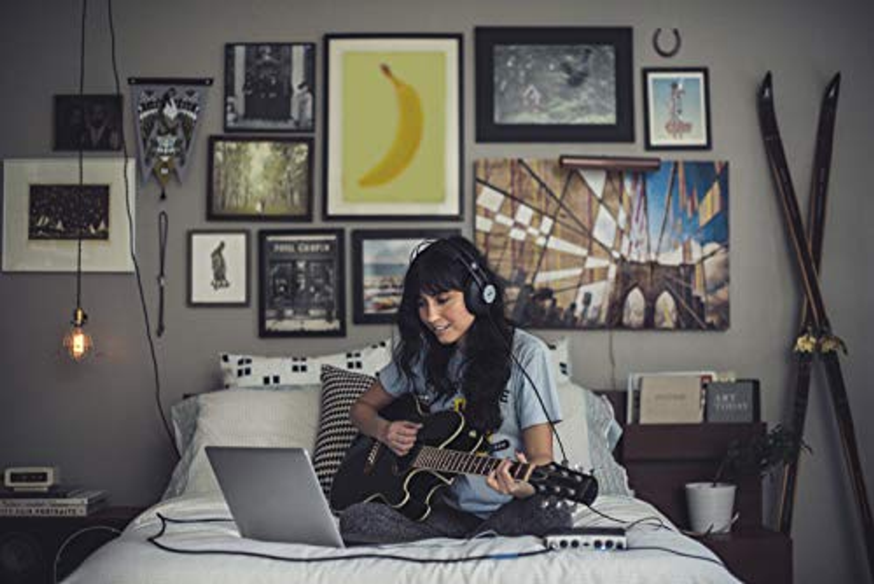 Donna che suona la chitarra sul letto, con laptop e cuffie.