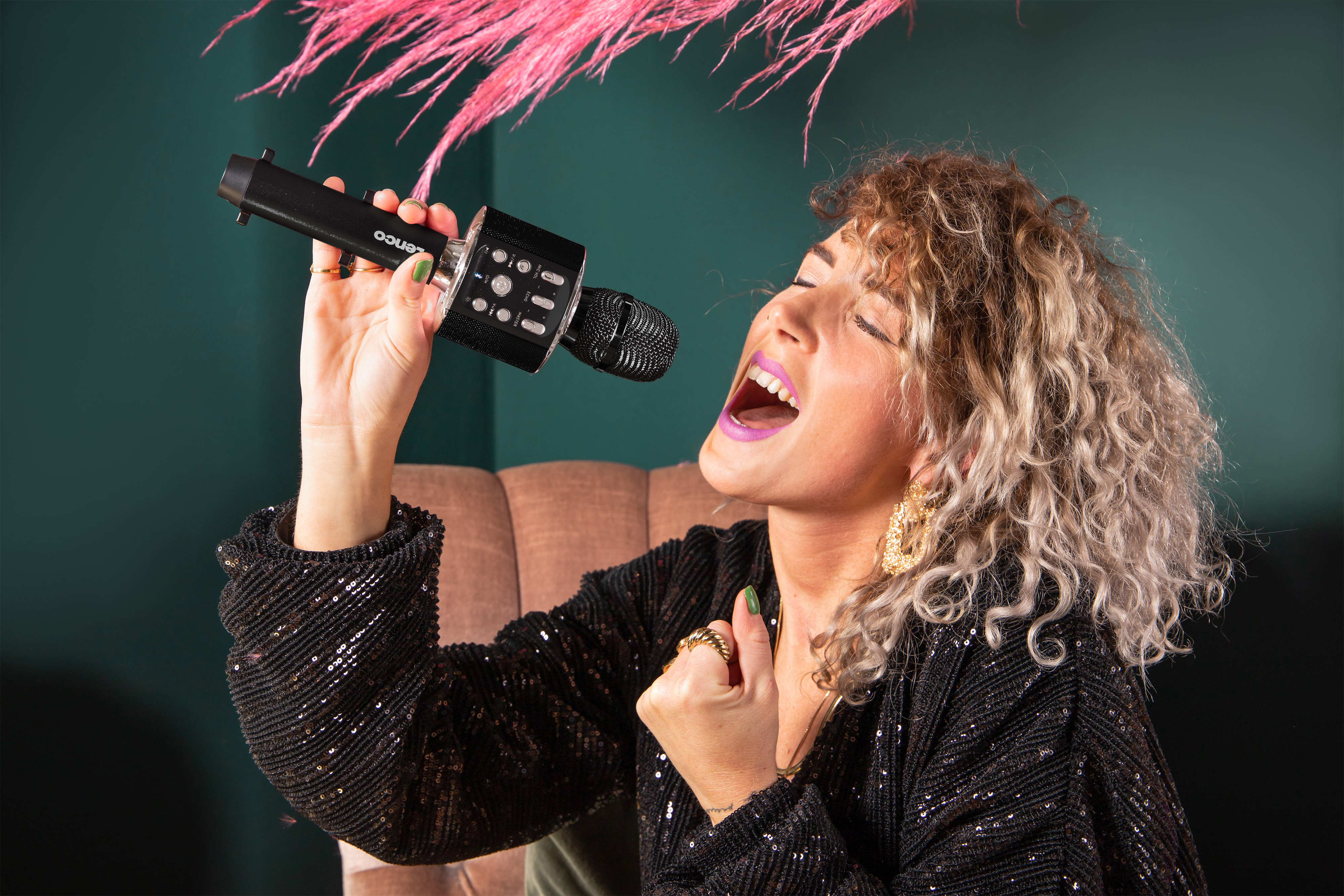 Een vrouw met krullend haar zingt in een microfoon. Roze bloemen staan achter haar.