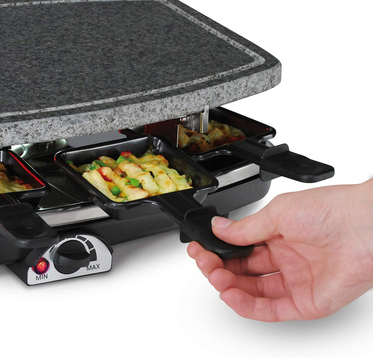Raclette-Grill mit Käse und Gemüse in den Schalen. Eine Hand zieht eine Schale mit Essen heraus.