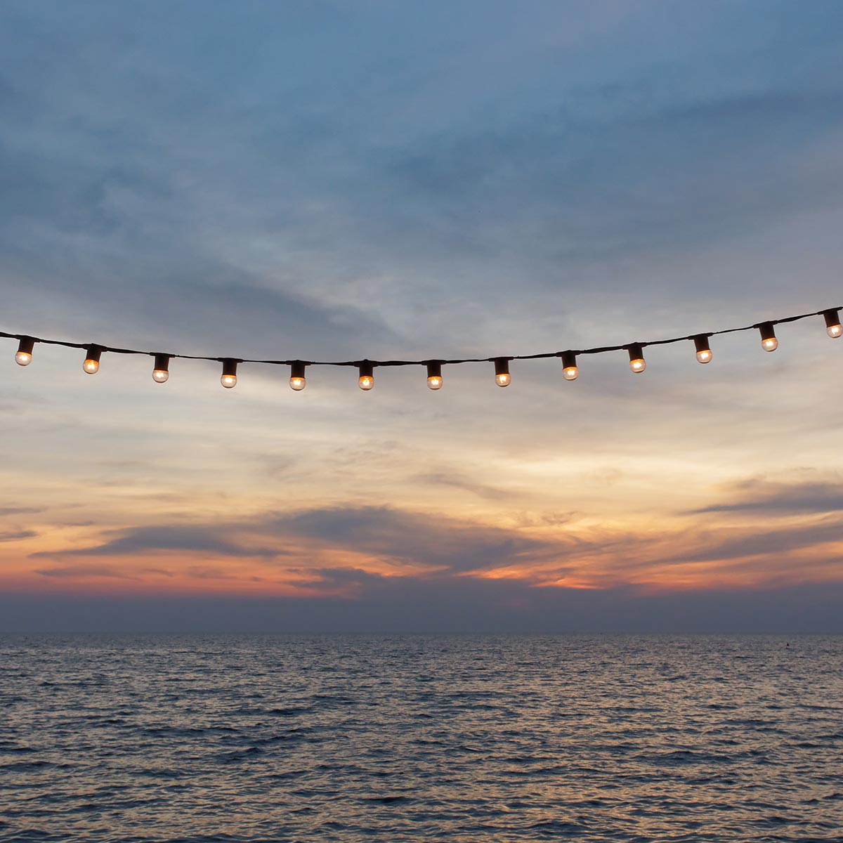 Eine Lichterkette vor einem Sonnenuntergang. Die Lichter sind rund und warmfarbig. Darunter das Meer.