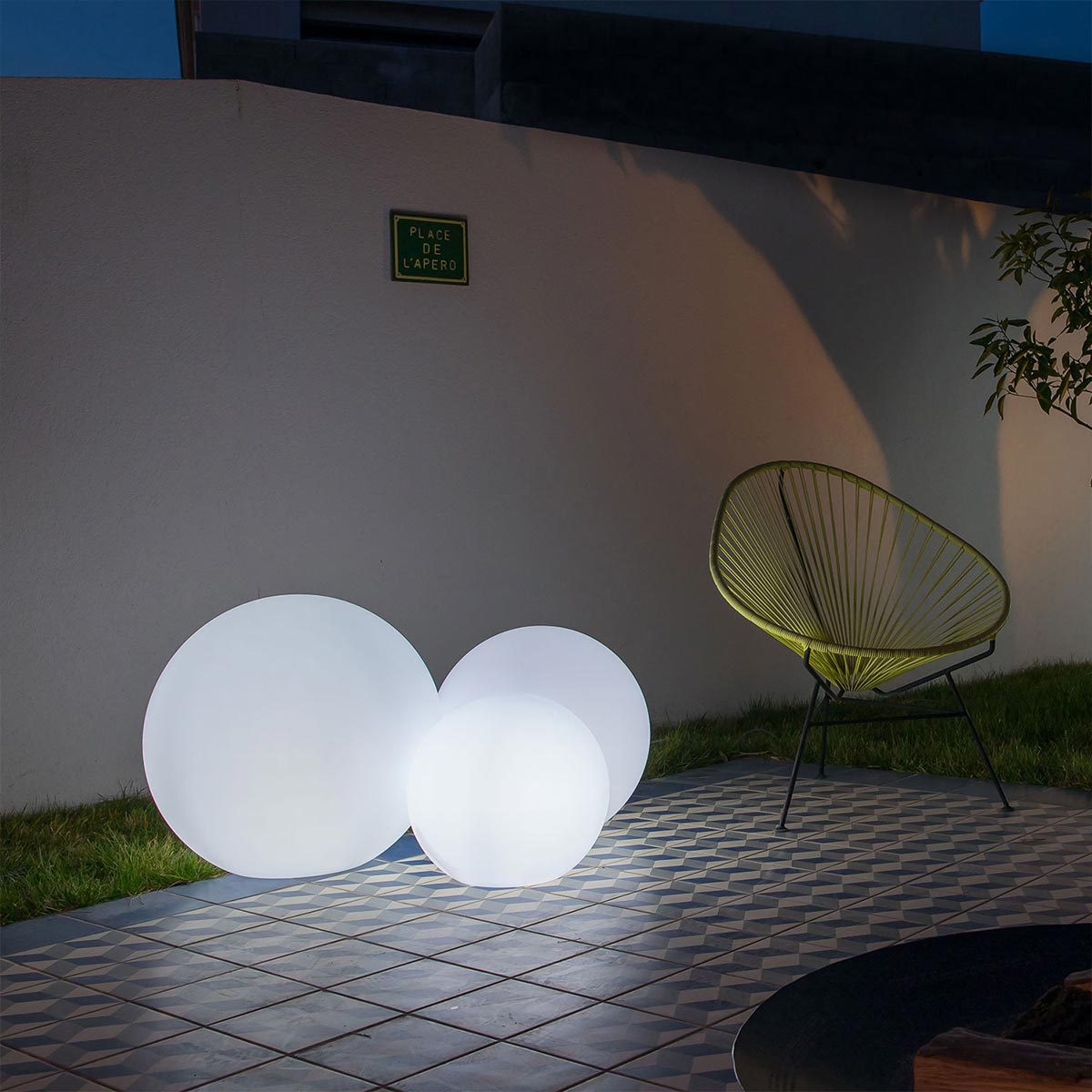 Drei beleuchtete weiße Kugeln draußen bei Nacht, mit einem grünen Stuhl, auf einer gefliesten Terrasse.