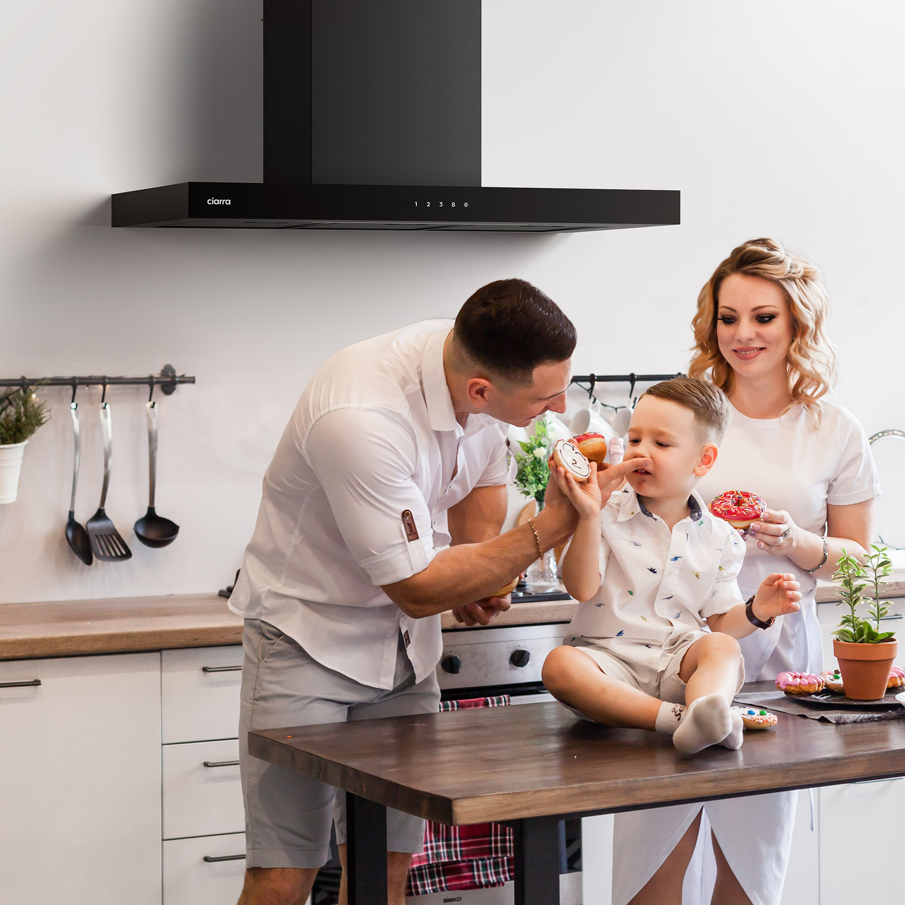 Una familia en una cocina, un hombre le da un donut a un niño mientras la mujer observa. Hay una campana extractora arriba.