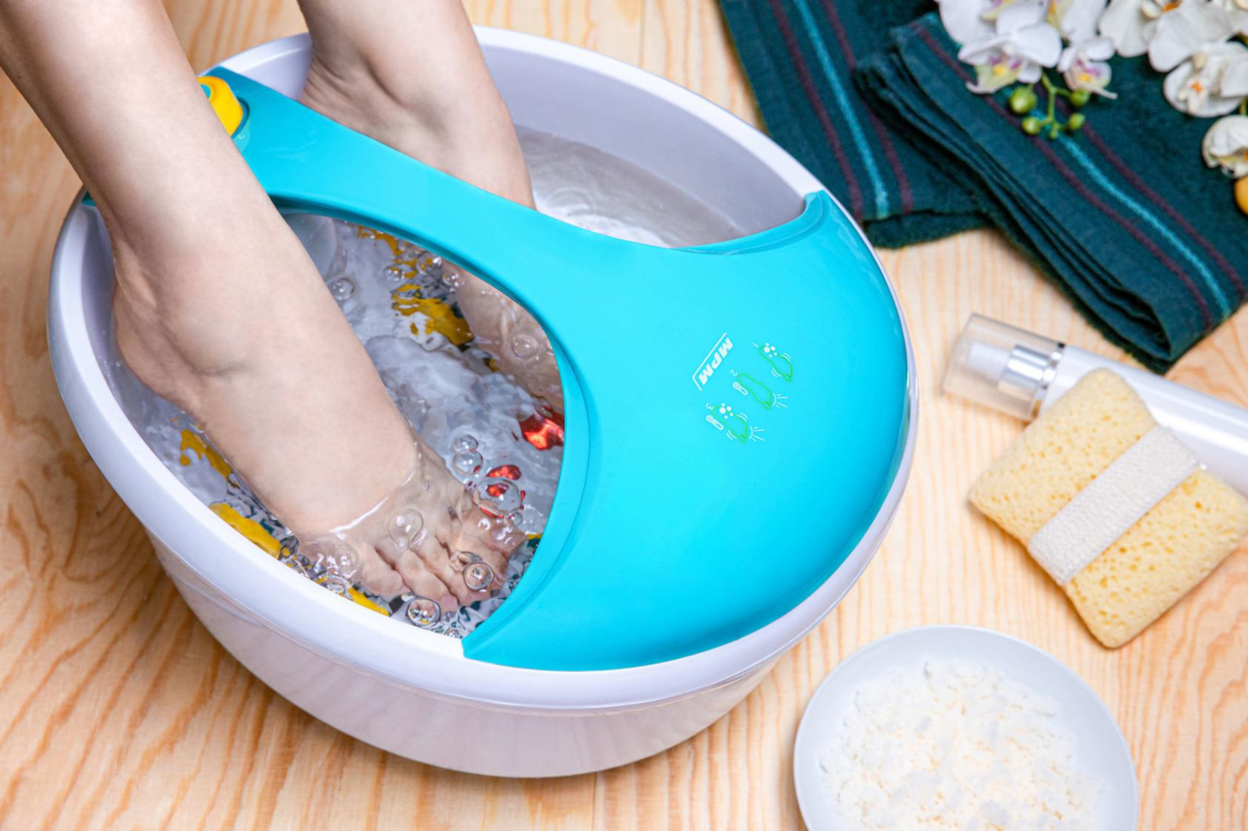 Pies en un baño de pies con agua. Baño turquesa. Cuenco blanco con agua y exfoliante al lado.
