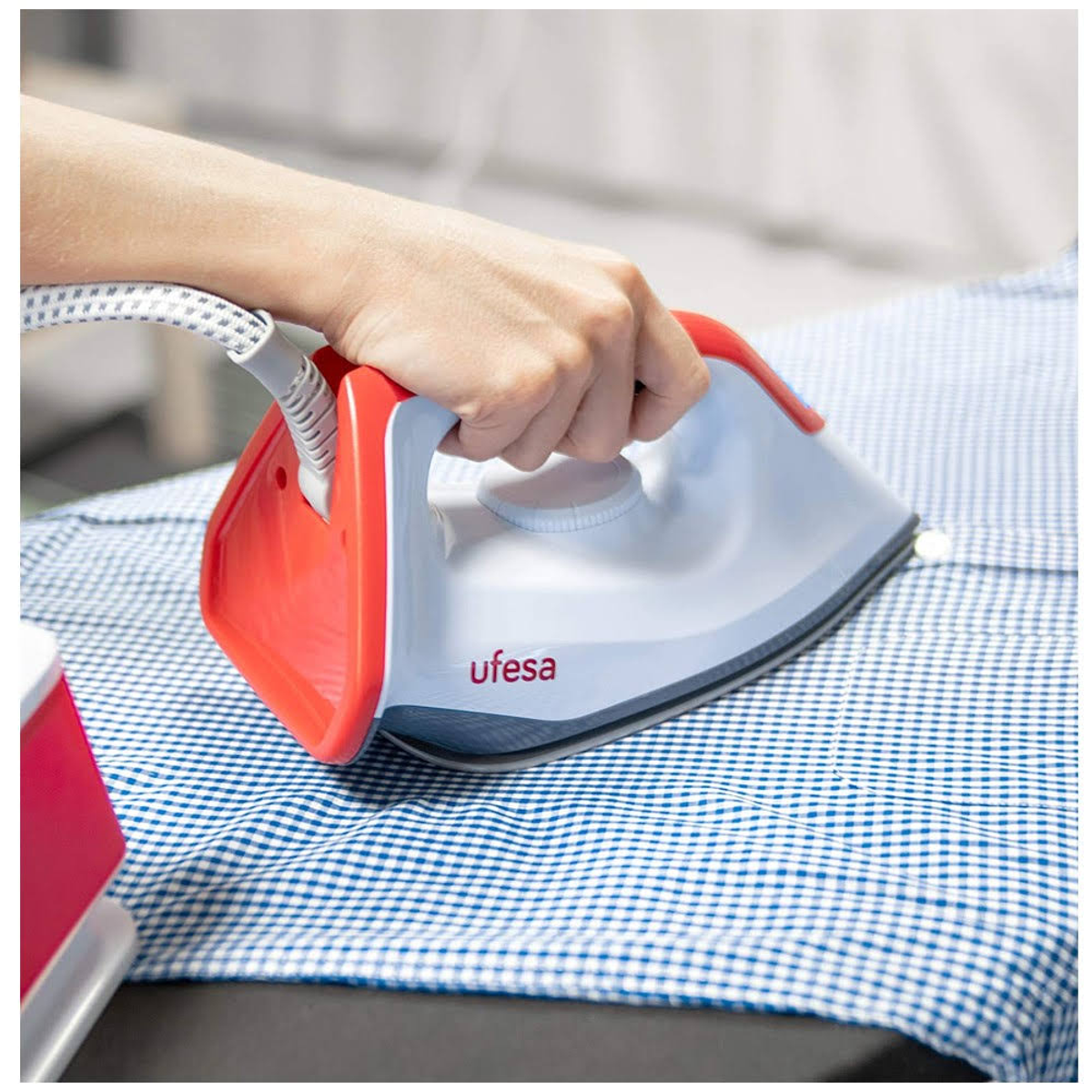 Una mano planchando una camisa a cuadros azul y blanco. La plancha es roja y blanca.