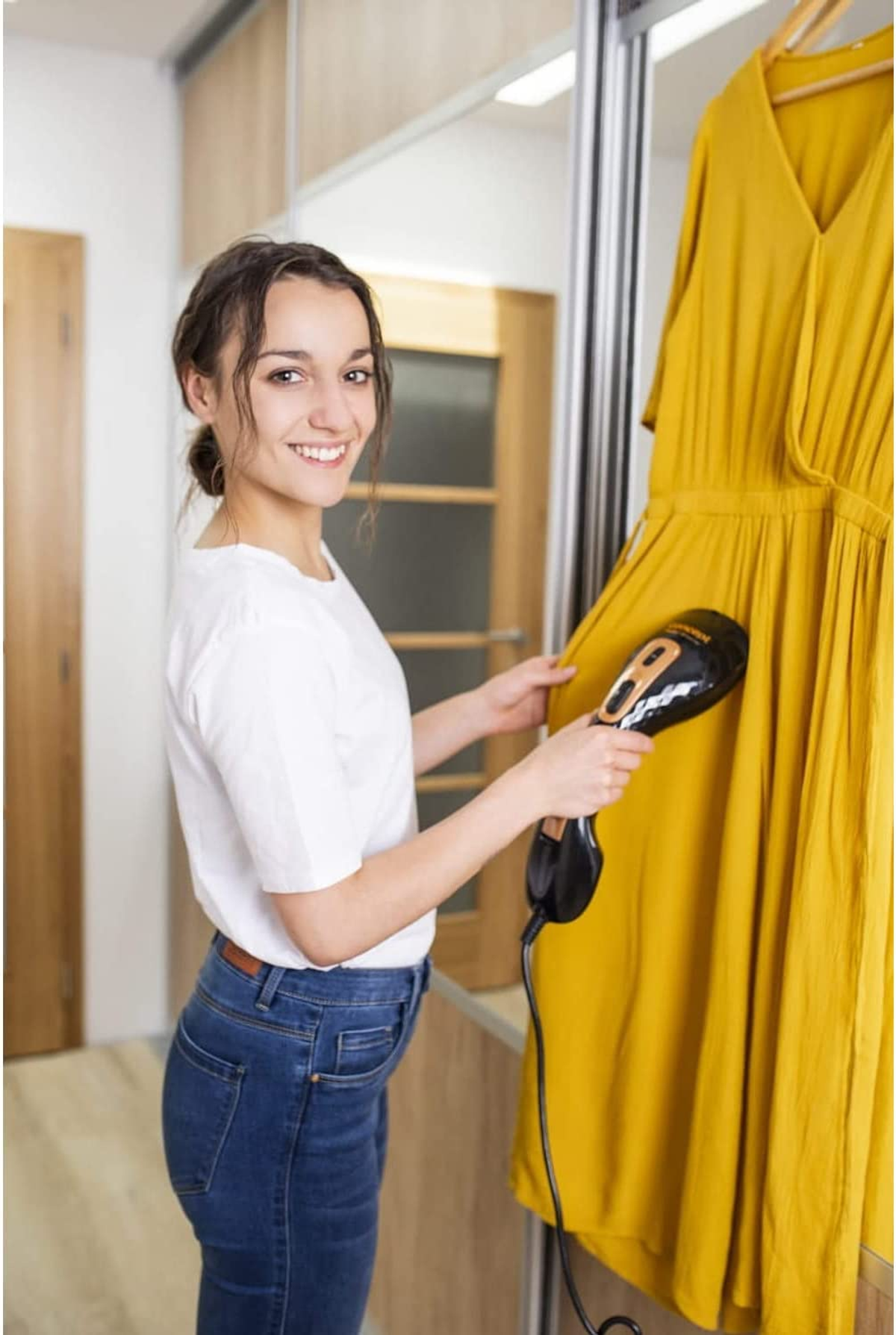 Una donna che stira a vapore un vestito giallo appeso a un'asta dell'armadio, sorridendo alla fotocamera.