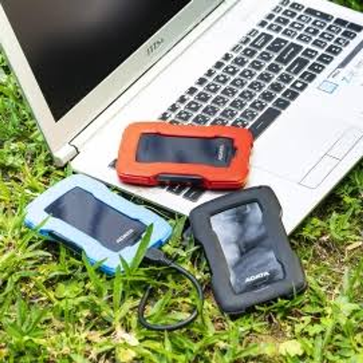 Laptop z trzema zewnętrznymi dyskami twardymi w kolorze czerwonym, niebieskim i czarnym, leżącymi na trawie.