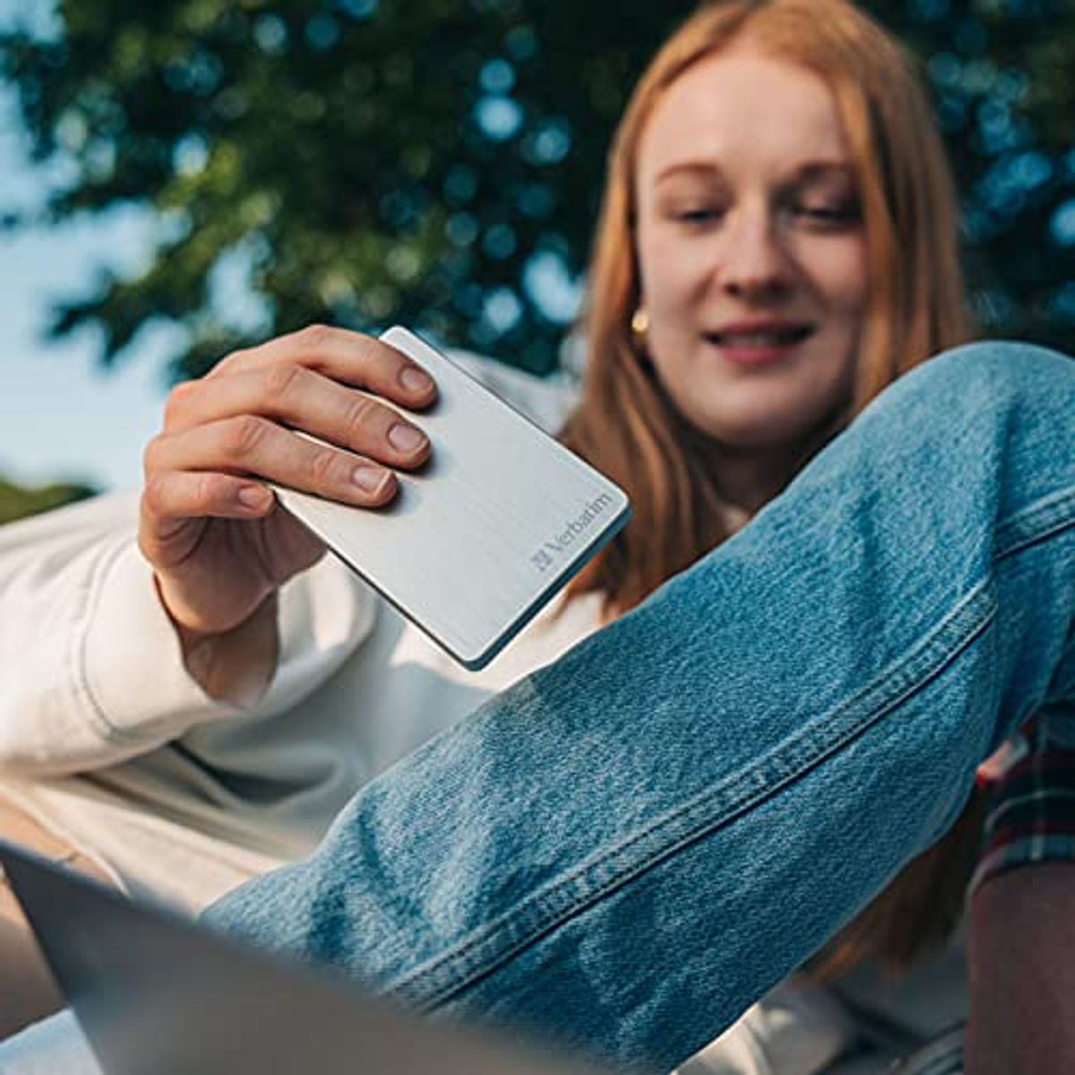 Kobieta trzymająca srebrny zewnętrzny dysk twardy, siedząc na zewnątrz.