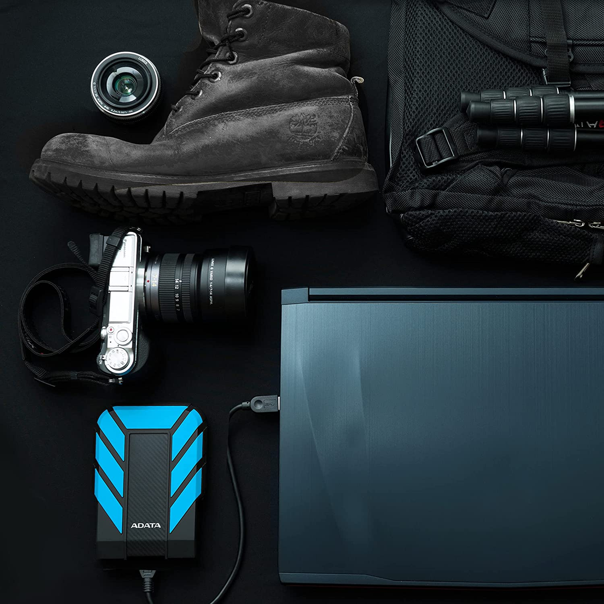 Czarny stół z czarnym plecakiem, aparatem, butem, obiektywami, laptopem i czarnym dyskiem twardym.