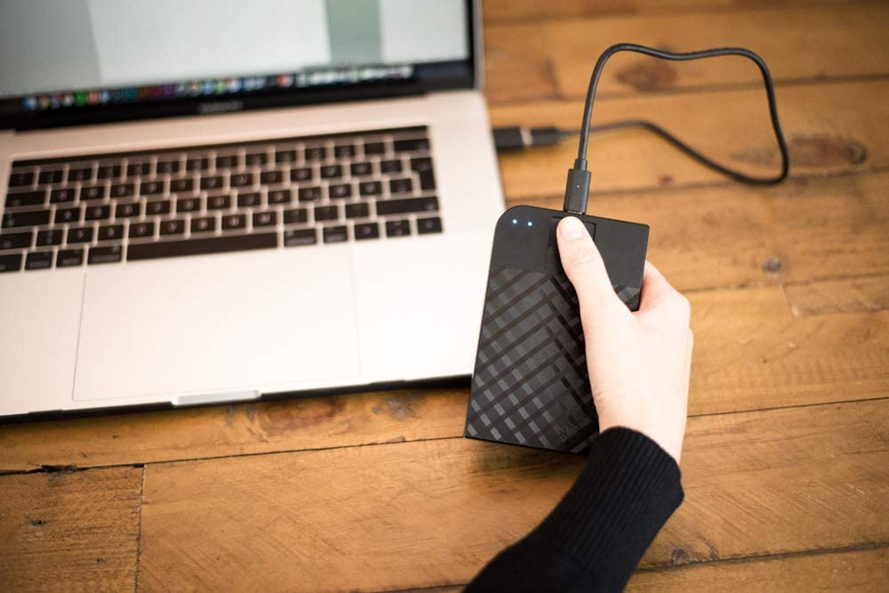Dłoń trzymająca czarny zewnętrzny dysk twardy podłączony do laptopa.