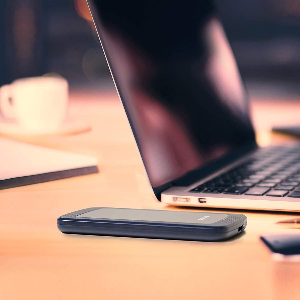 Laptop z czarnym zewnętrznym dyskiem twardym i kubkiem na drewnianym biurku.