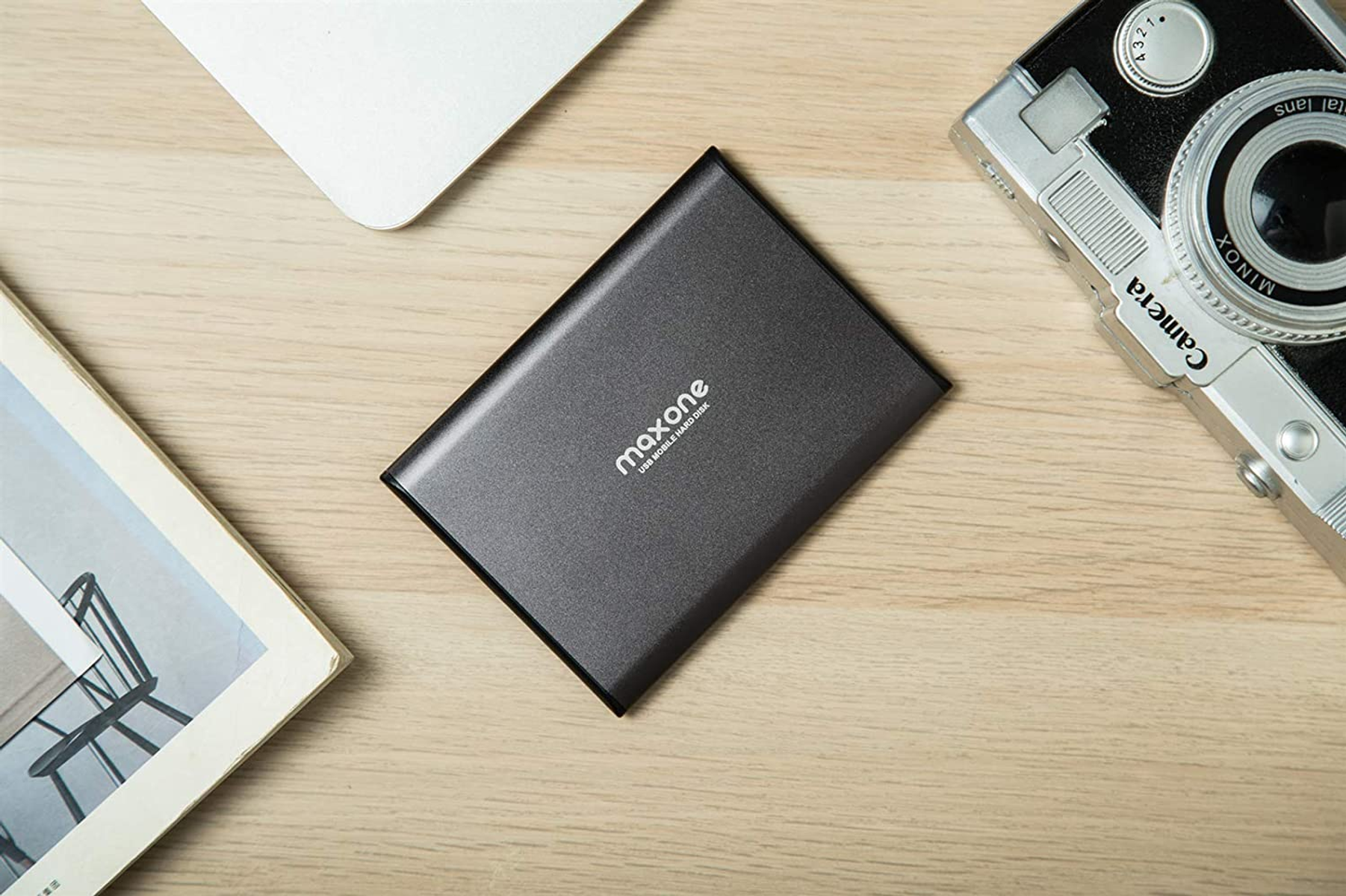 Un disco rigido grigio scuro su una superficie di legno con una fotocamera e un libro.