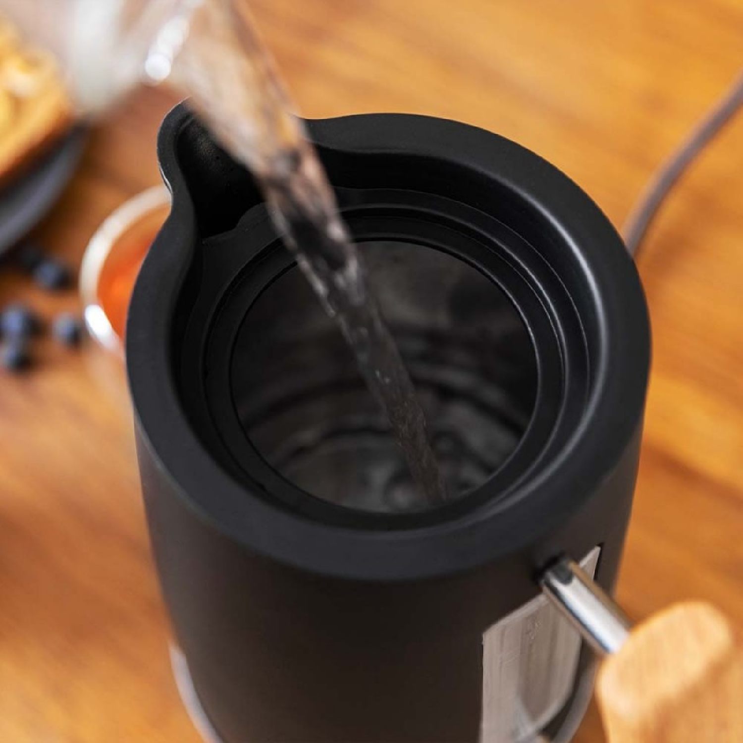 Woda wlewana do czarnego czajnika elektrycznego. Drewniany stół, rozmyte tło.