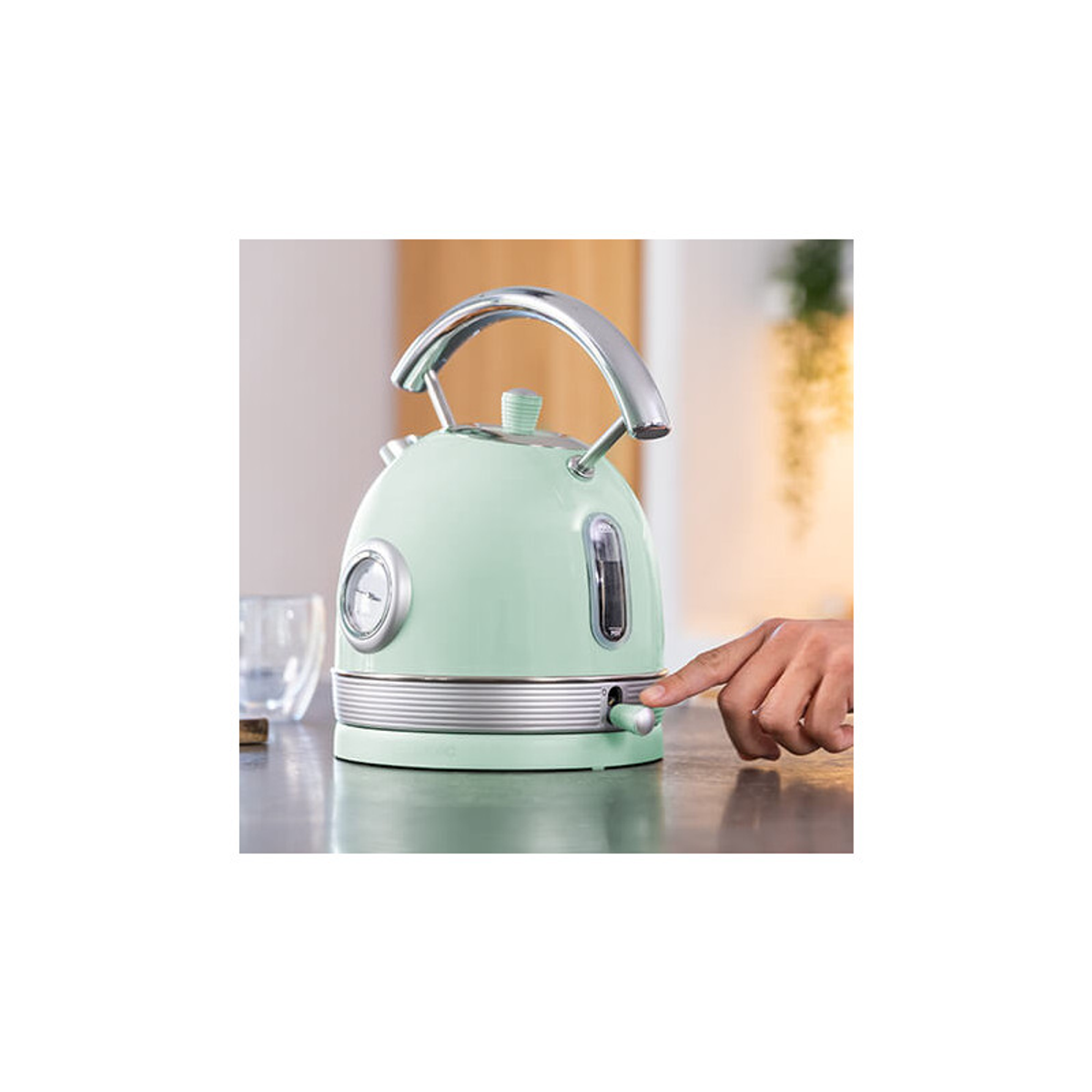 Ręka osoby włącza miętowo-zielony czajnik ze srebrnymi akcentami; widoczny jest szklany kubek i tło.