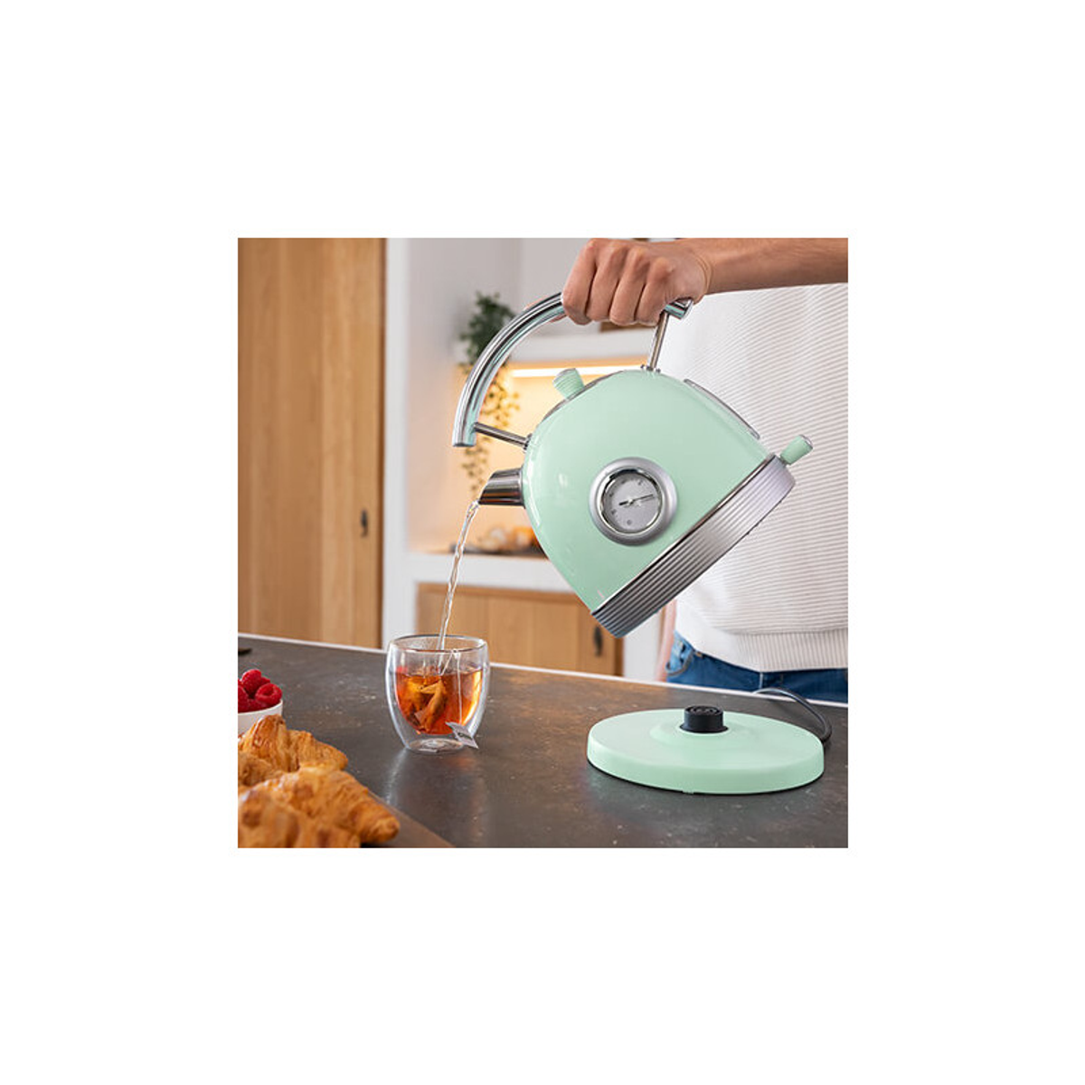 Osoba nalewa gorącą wodę z miętowego czajnika do szklanki. Widoczne rogaliki i maliny.