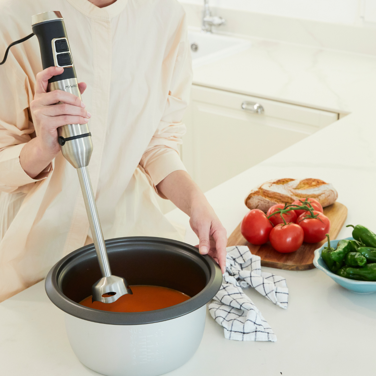 Persona che usa un frullatore a immersione in una pentola di zuppa. Pomodori e peperoni a lato.