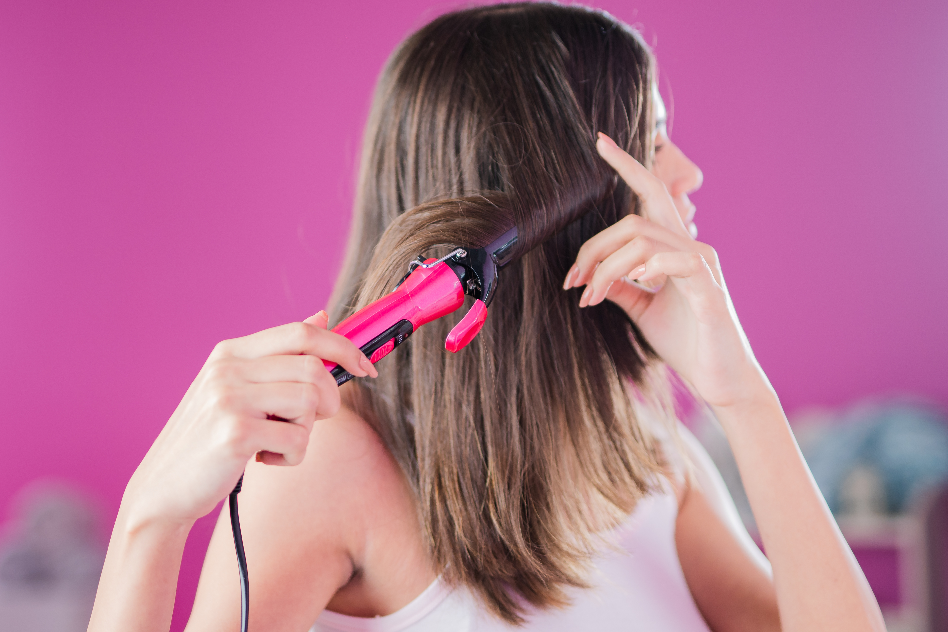 Una mujer con cabello castaño se riza el cabello con una plancha rosa. Fondo rosa.