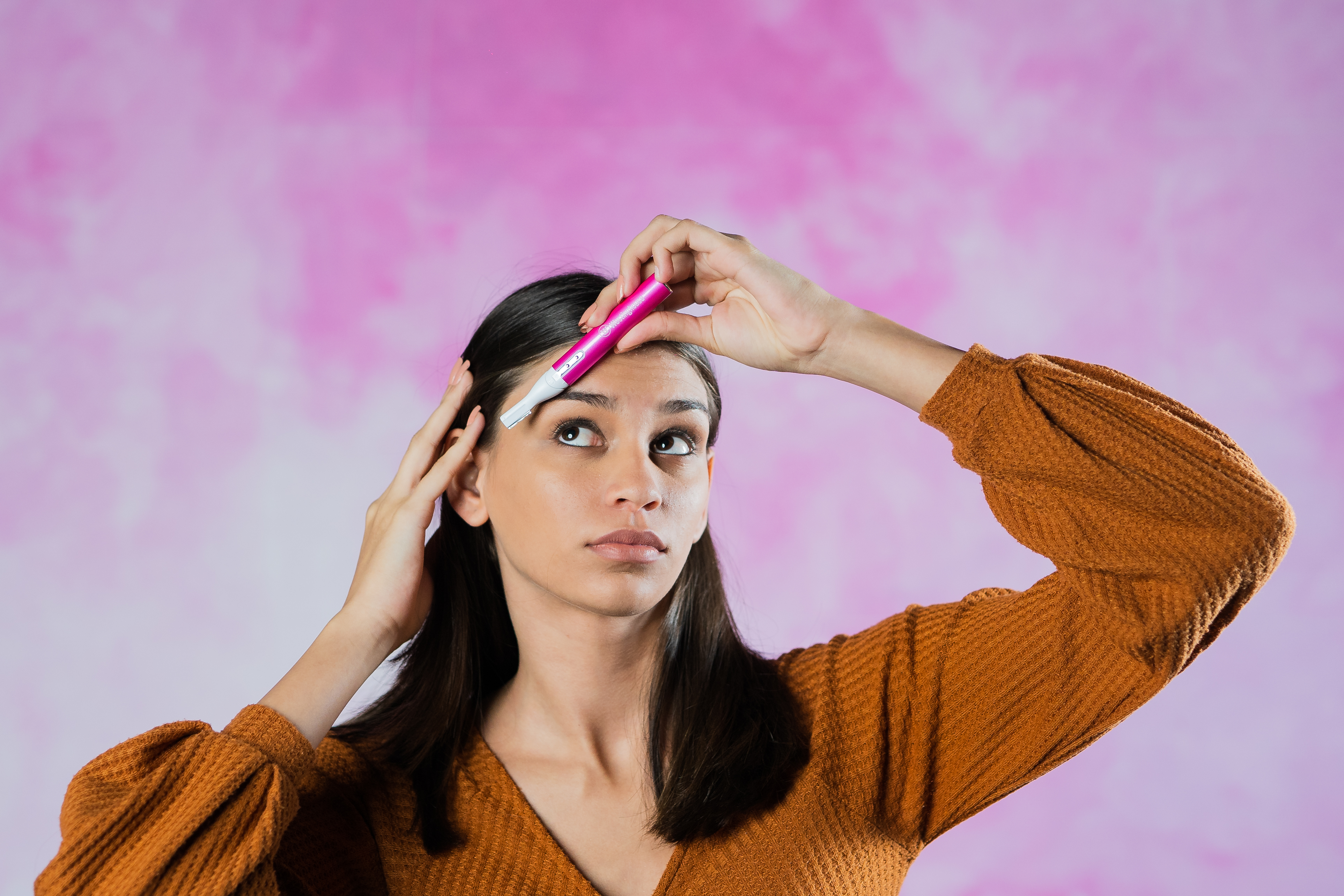 Donna con camicia arancione tocca la fronte con un rifinitore per sopracciglia rosa, su sfondo rosa.