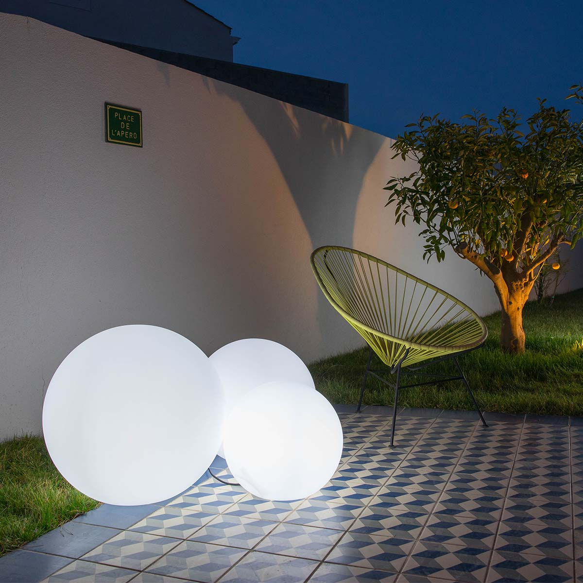 Beleuchtete Außenszene. Drei leuchtende, kugelförmige Lichter auf einer gefliesten Terrasse, mit grünem Stuhl.