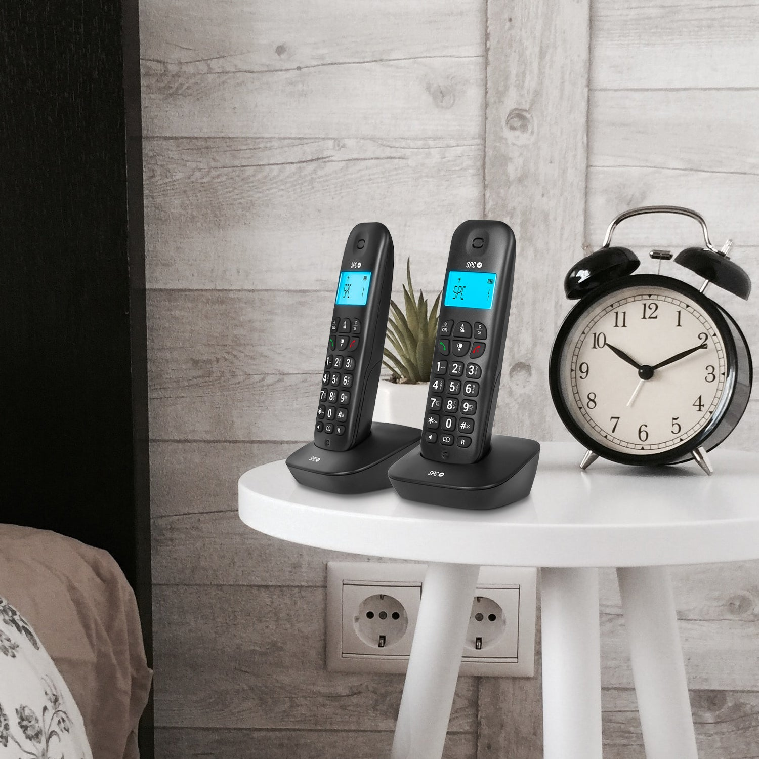 Due telefoni cordless neri su un tavolo bianco, con un orologio e una pianta sullo sfondo.