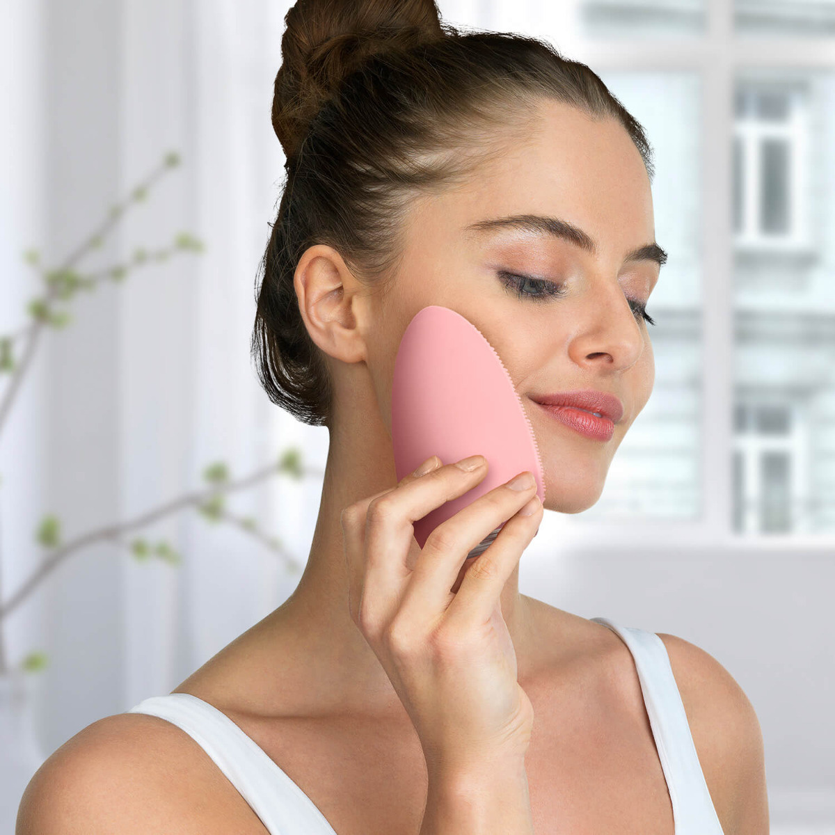 Eine Frau benutzt eine rosa Gesichtsreinigungsbürste auf ihrer Wange. Weißer Hintergrund.