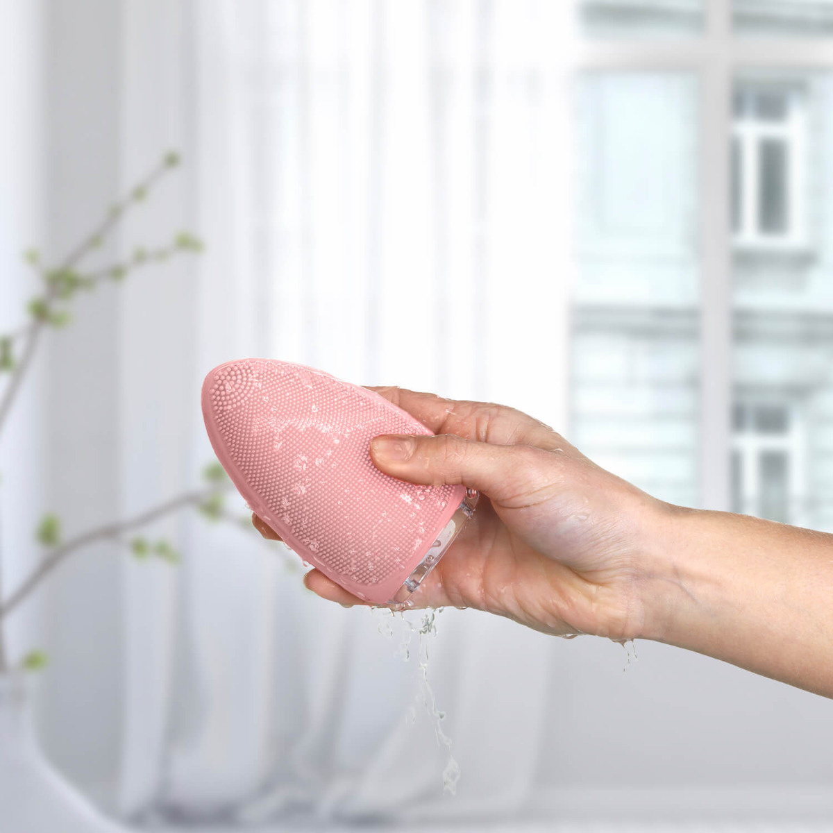 Hand hält rosa Silikon-Gesichtsbürste, Wasser läuft davon. Weißer Hintergrund.