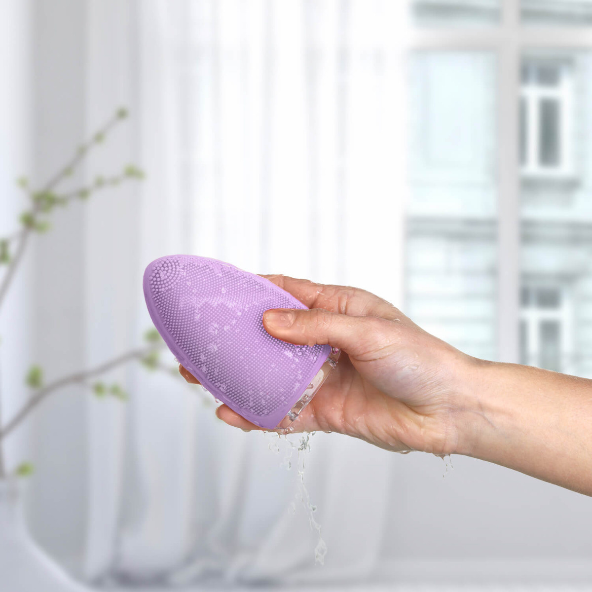 Eine Hand hält eine lila Gesichtsreinigungsbürste, Wasser tropft davon. Weißer Hintergrund.