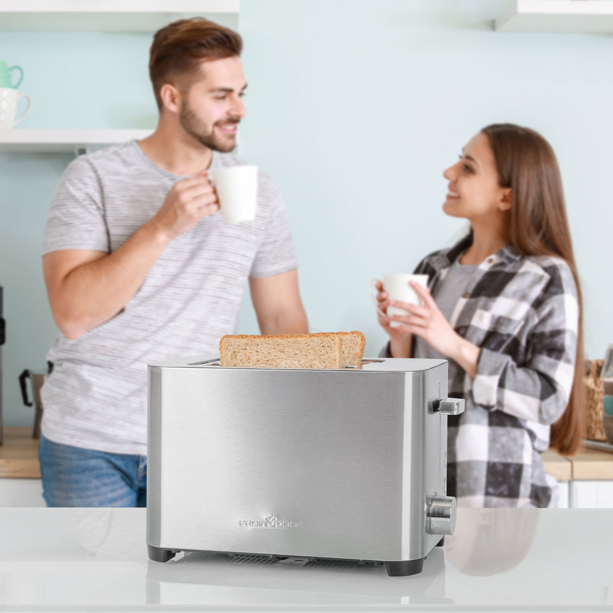 Ein Toaster aus Edelstahl mit Toast steht auf einem weißen Tisch. Ein Paar ist im unscharfen Hintergrund.