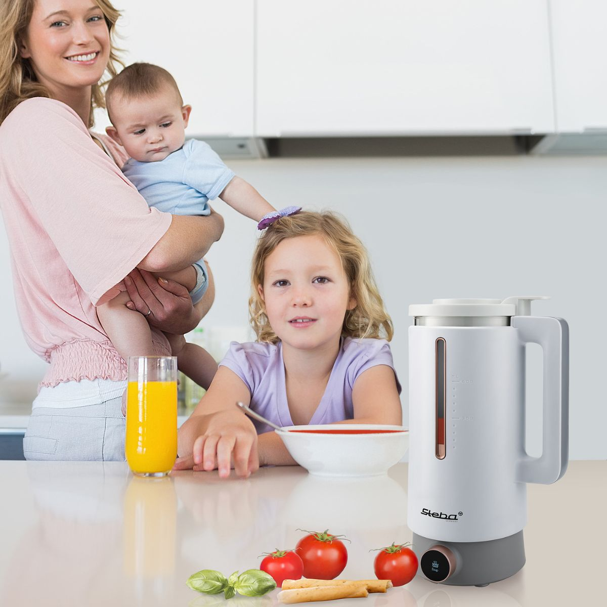 Familie am Tisch. Mutter hält Baby, Mädchen lächelt. Mixer und Zutaten auf dem Tisch.
