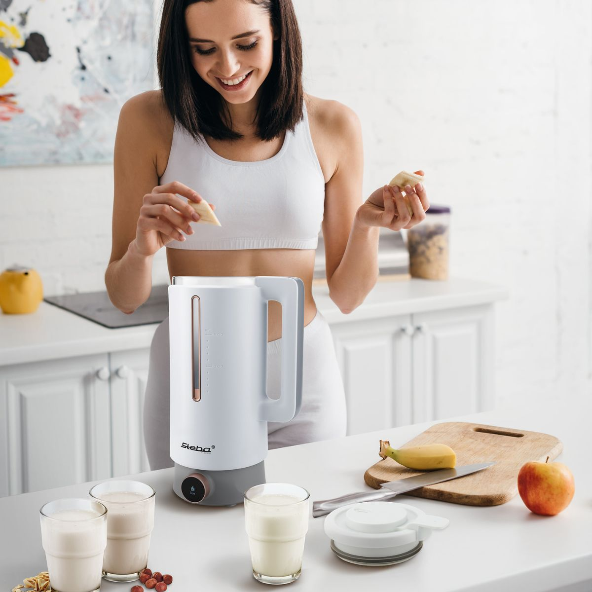 Frau in einer Küche bereitet Essen zu. Mixer, Obst und Gläser mit Milch sind auf der Theke zu sehen.