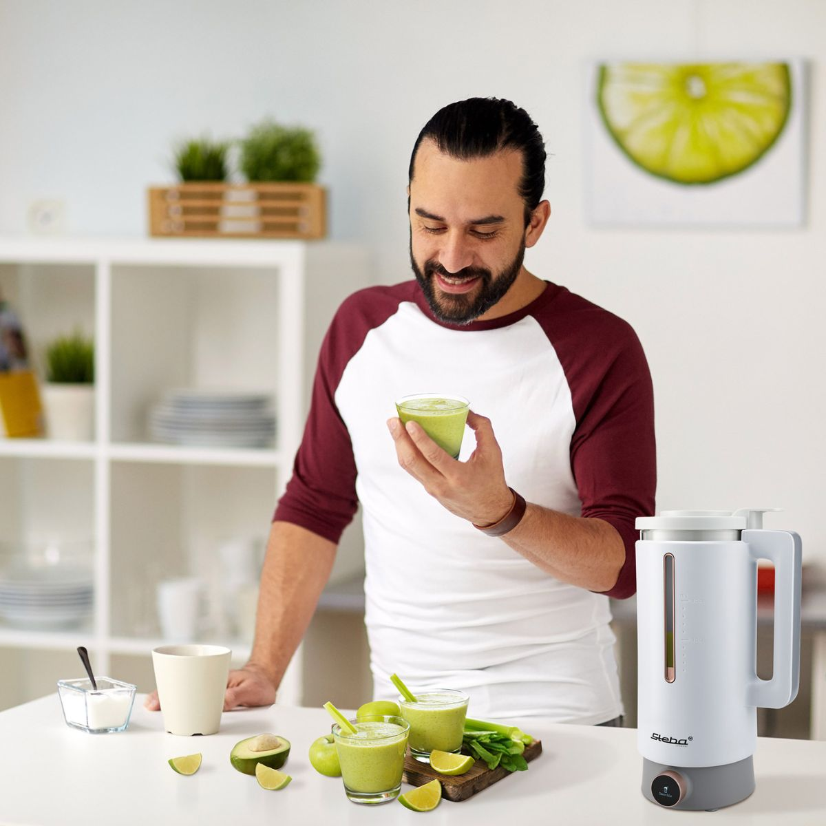 Uomo che tiene una bevanda verde, con ingredienti e un frullatore sul tavolo.
