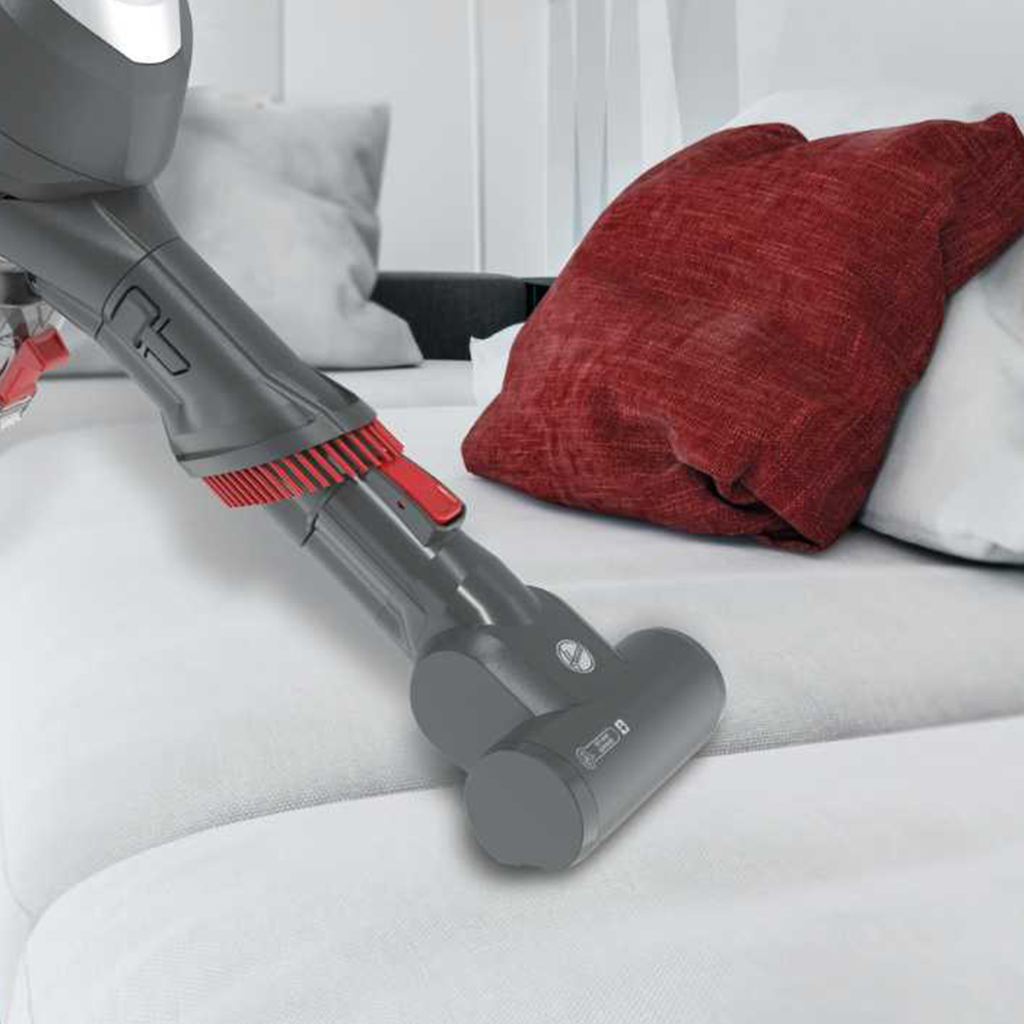 Ein grauer Staubsauger reinigt ein weißes Sofa. Auf dem Sofa liegt ein rotes Kissen.