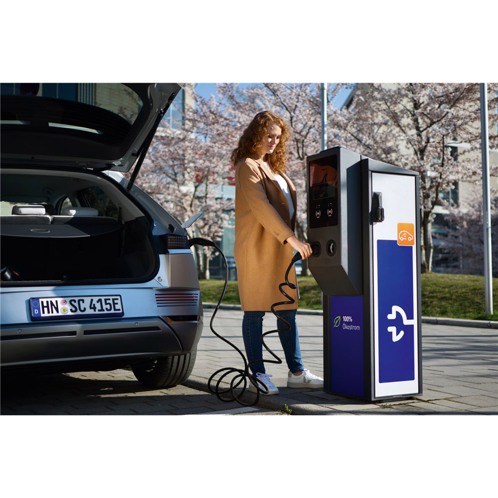 Donna che carica auto elettrica alla stazione. Auto argento, stazione blu e bianca.