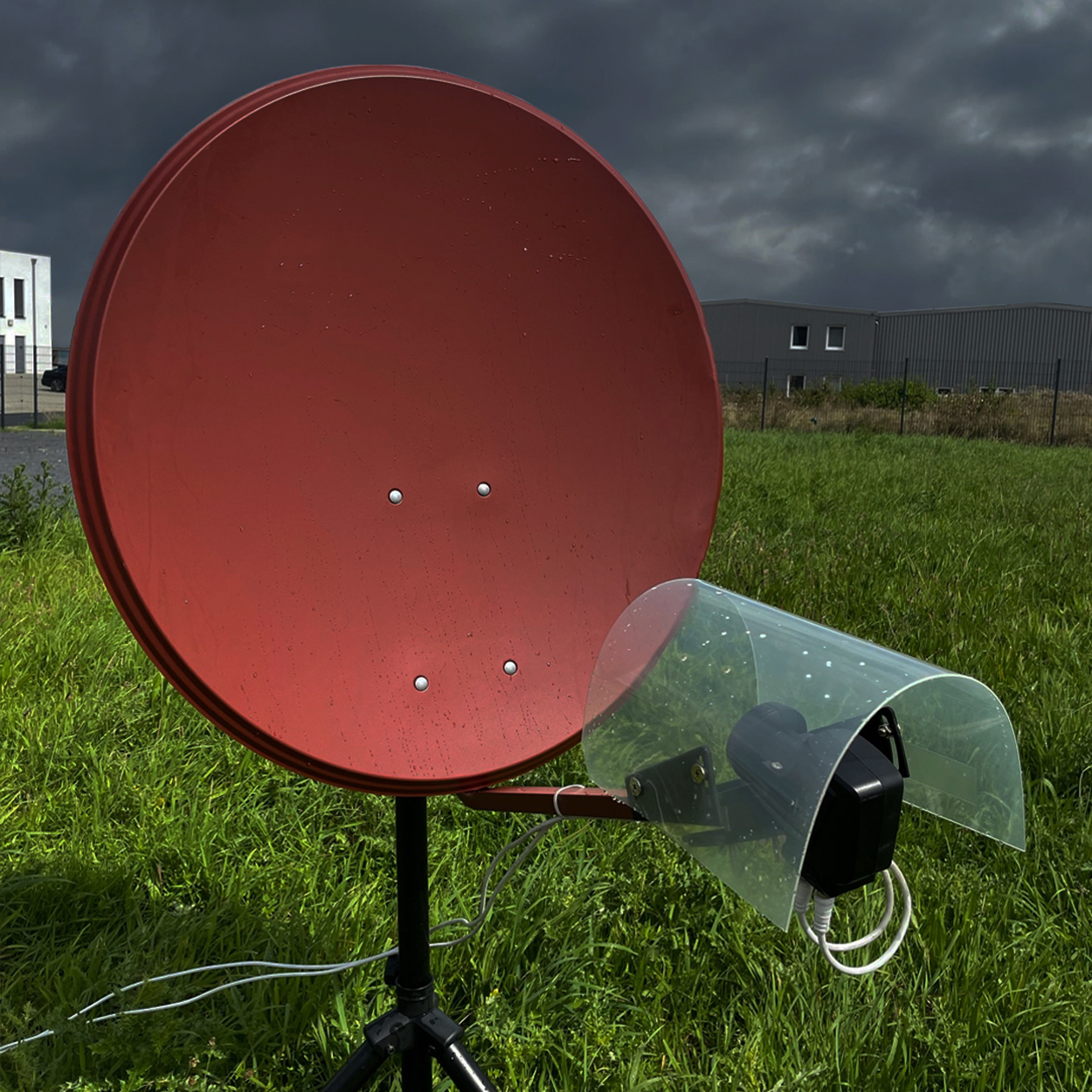 Roter Satellitenteller auf einem Stativ in einem grasbewachsenen Feld mit dunklem Himmel im Hintergrund.