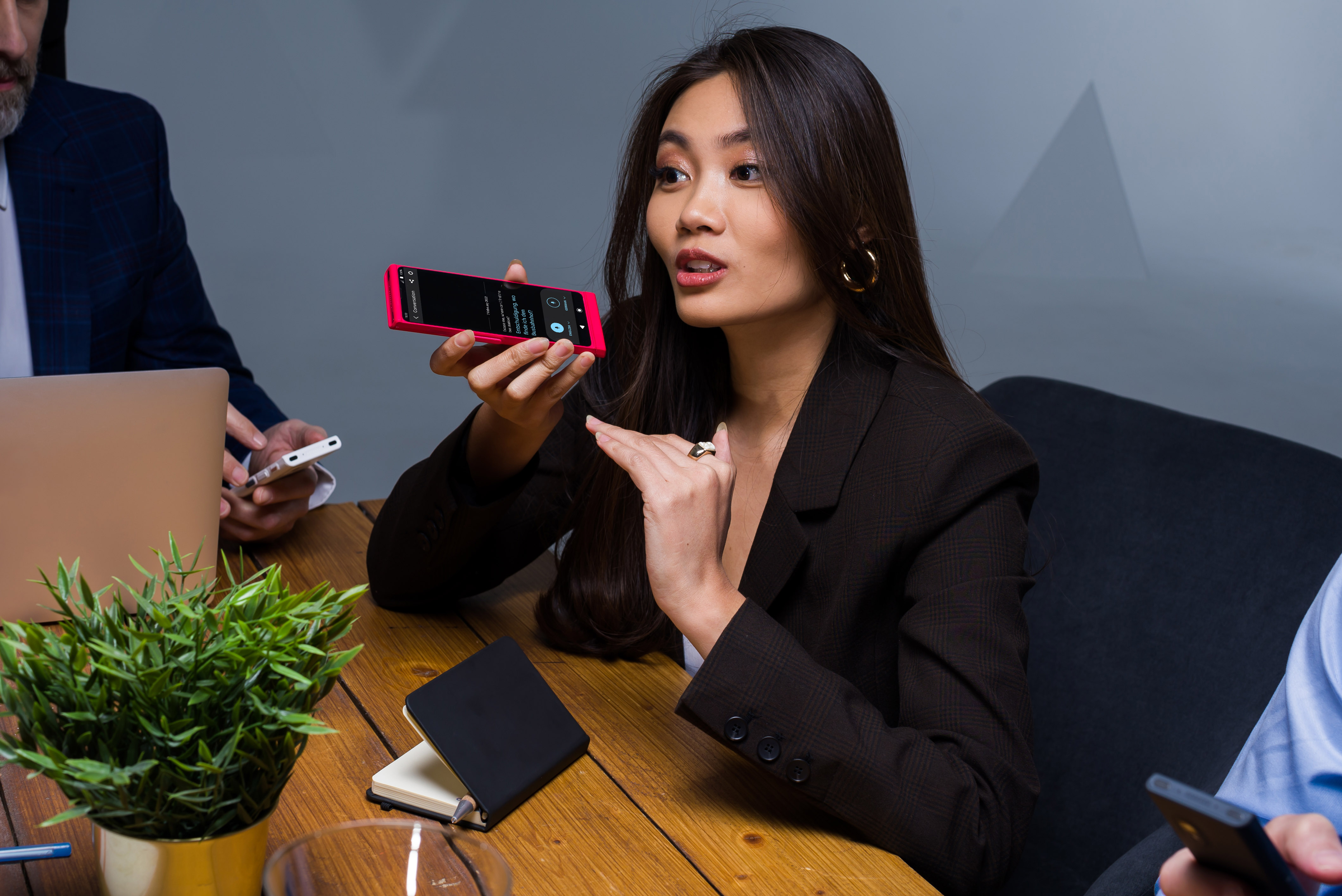 Eine Frau spricht mit einem rosa Telefon, im Hintergrund Kollegen und ein Laptop.