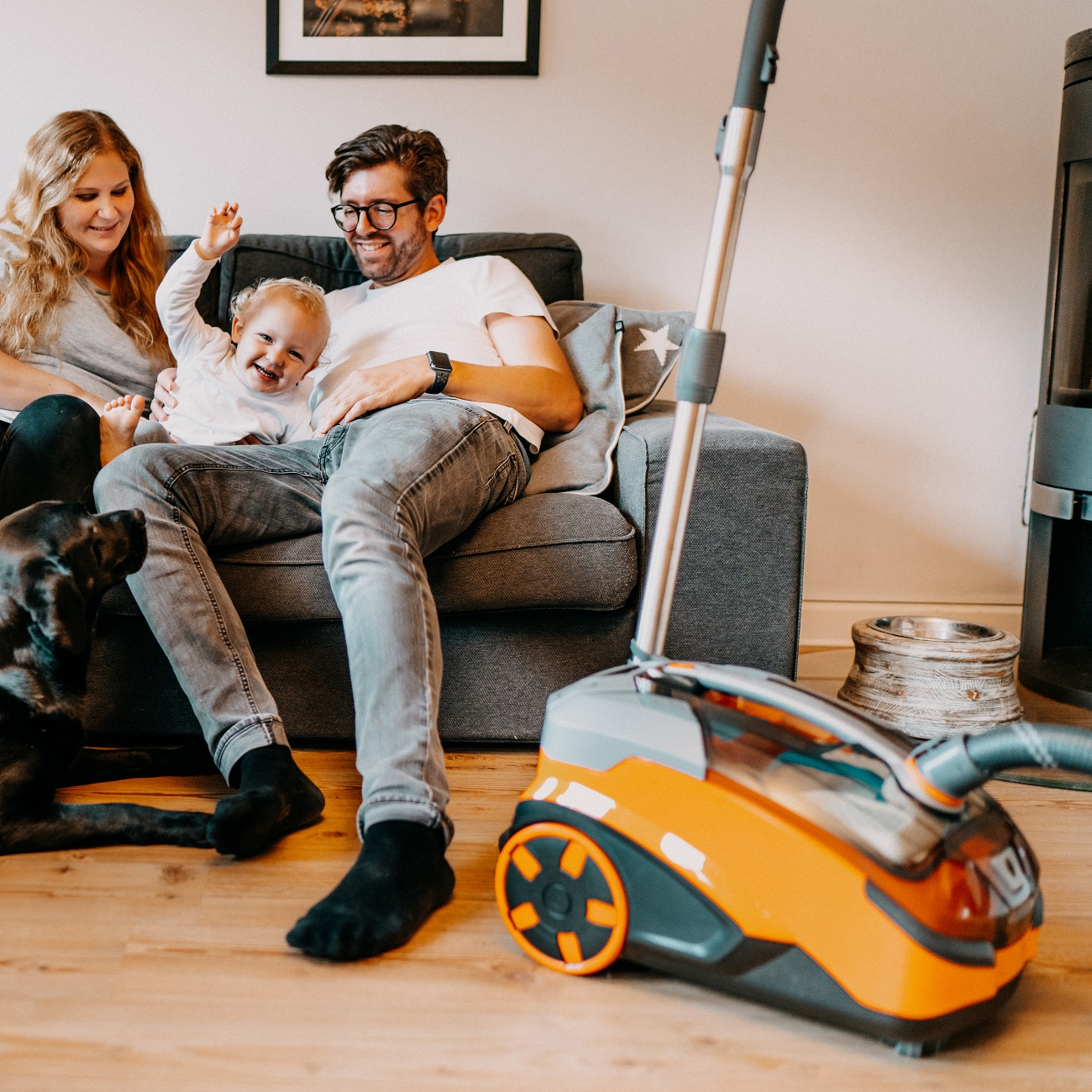 Eine Familie mit Baby und Hund im Wohnzimmer und ein Staubsauger.