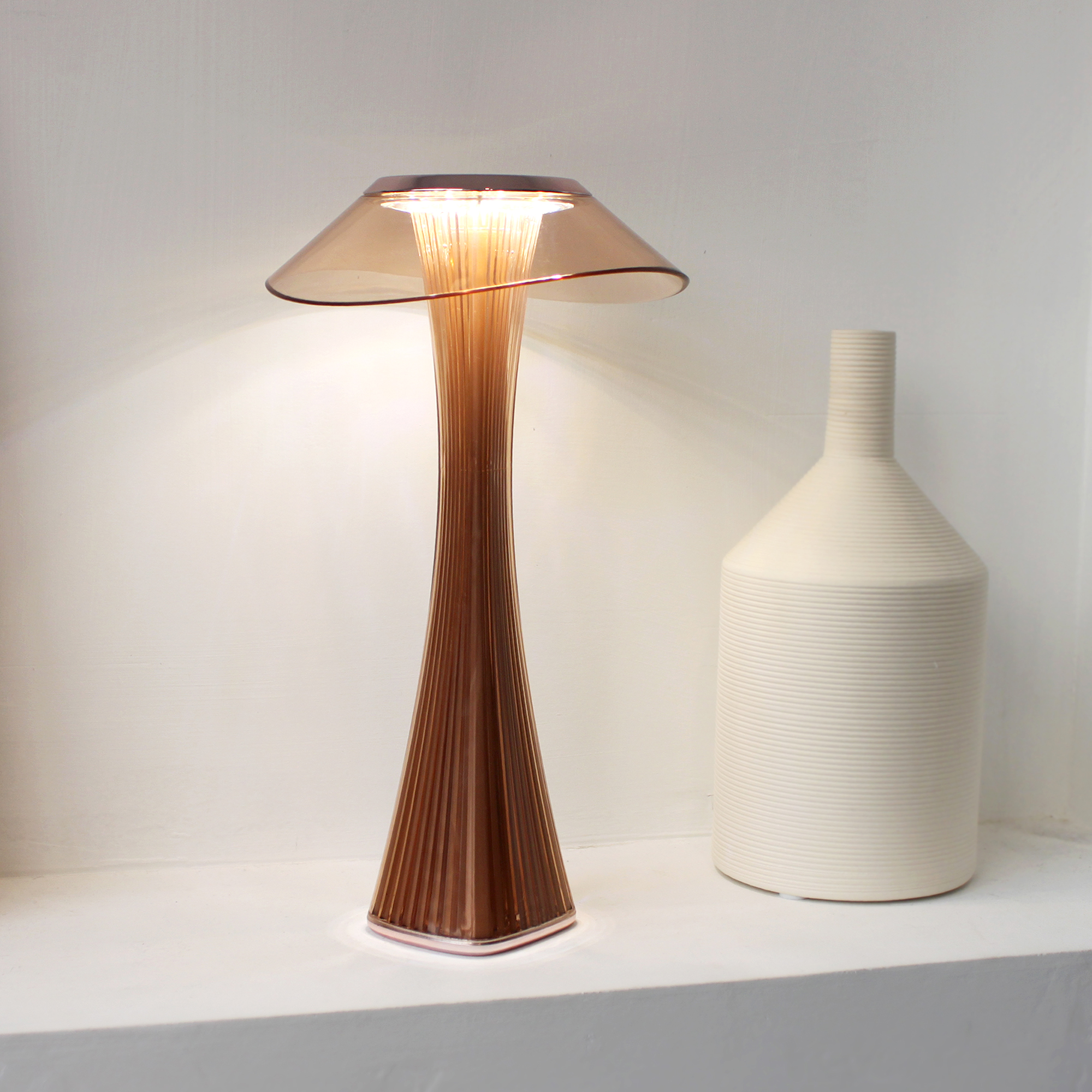Eine braune und klare Lampe auf einem weißen Regal, mit einer weißen Vase daneben.
