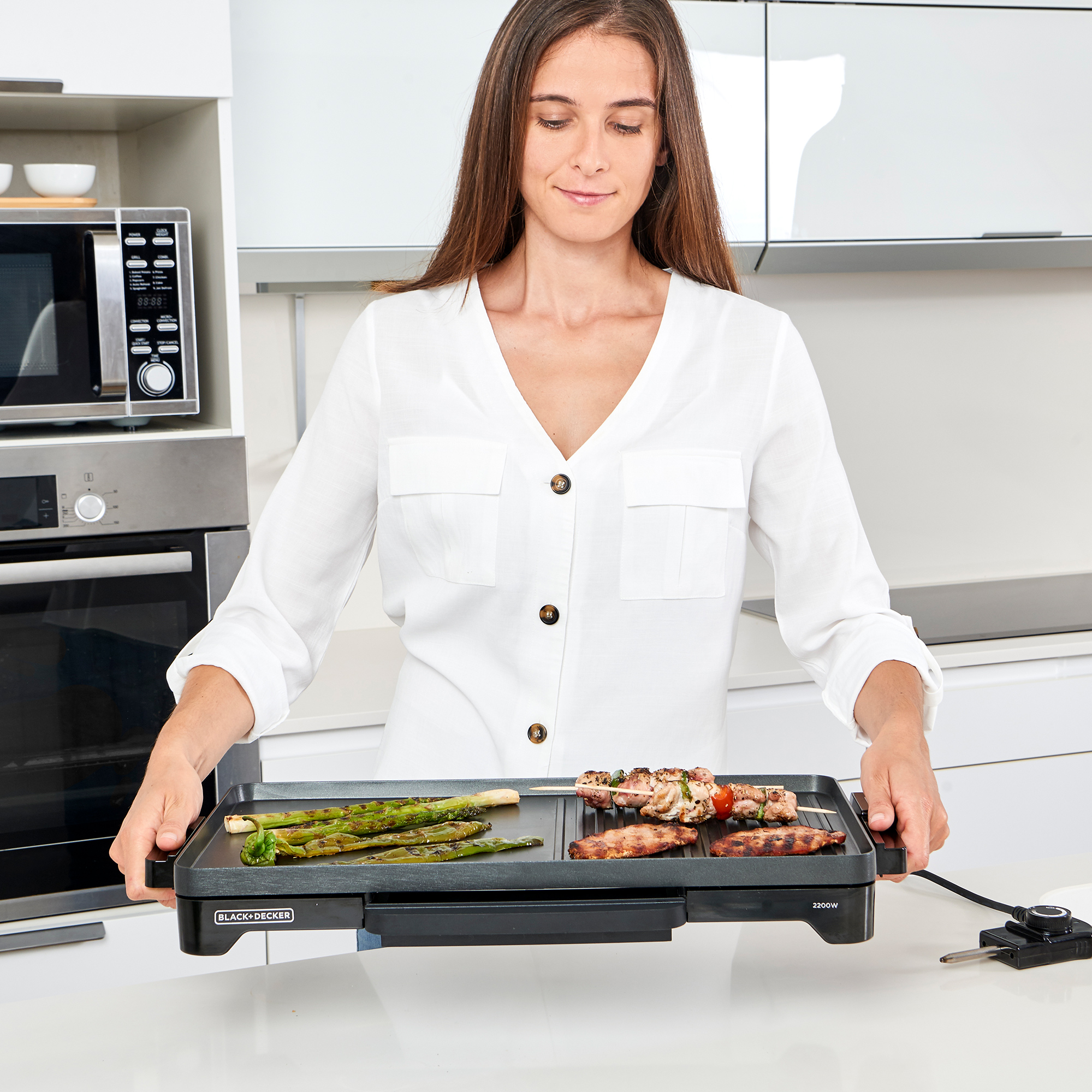 Kobieta trzymająca grilla Black+Decker z grillowanym jedzeniem. W tle kuchenka mikrofalowa.