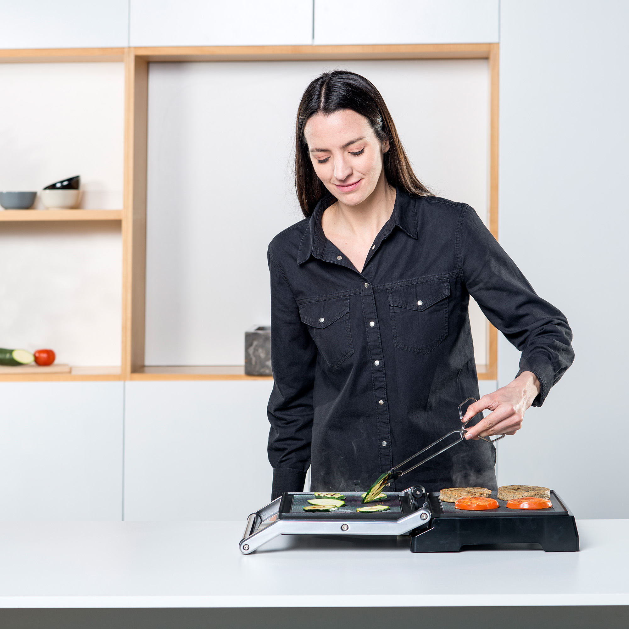 Kobieta gotuje na czarnym grillu elektrycznym. Używa szczypiec do obsługi jedzenia na białym blacie.