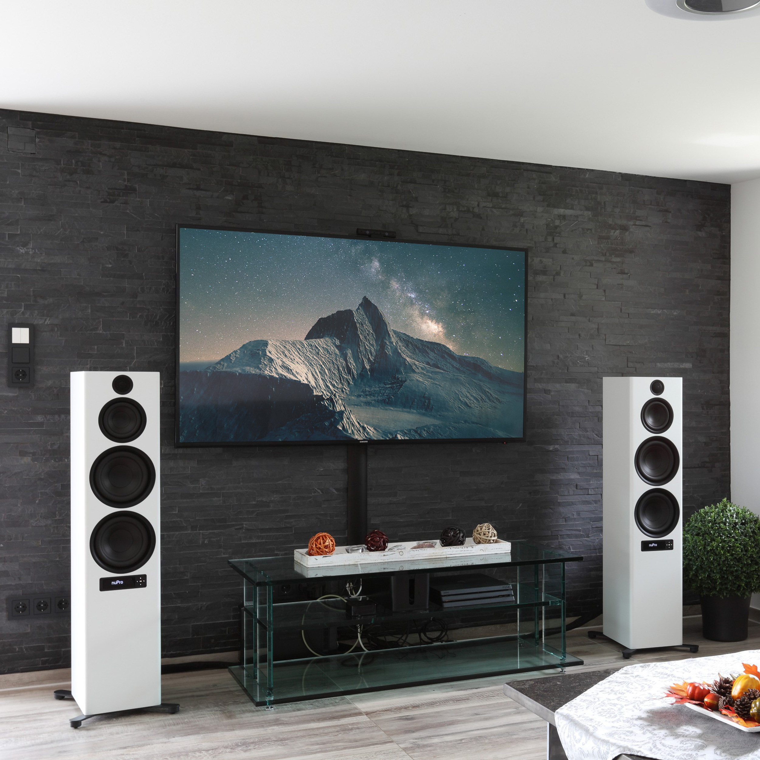 Ein Wohnzimmer mit einem Fernseher an einer schwarzen Backsteinmauer, weißen Lautsprechern, einem Glastisch und Dekorationsartikeln.