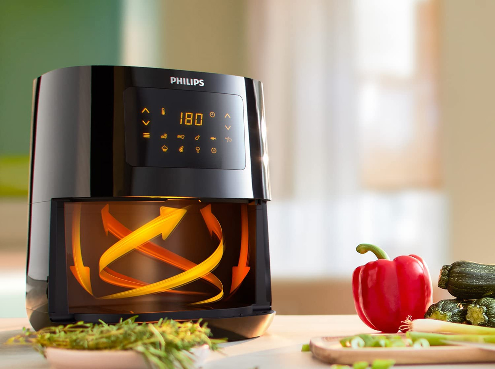 Schwarze Heißluftfritteuse mit Digitalanzeige. Rote Paprika, Zucchini und Kräuter daneben auf einem Tisch.