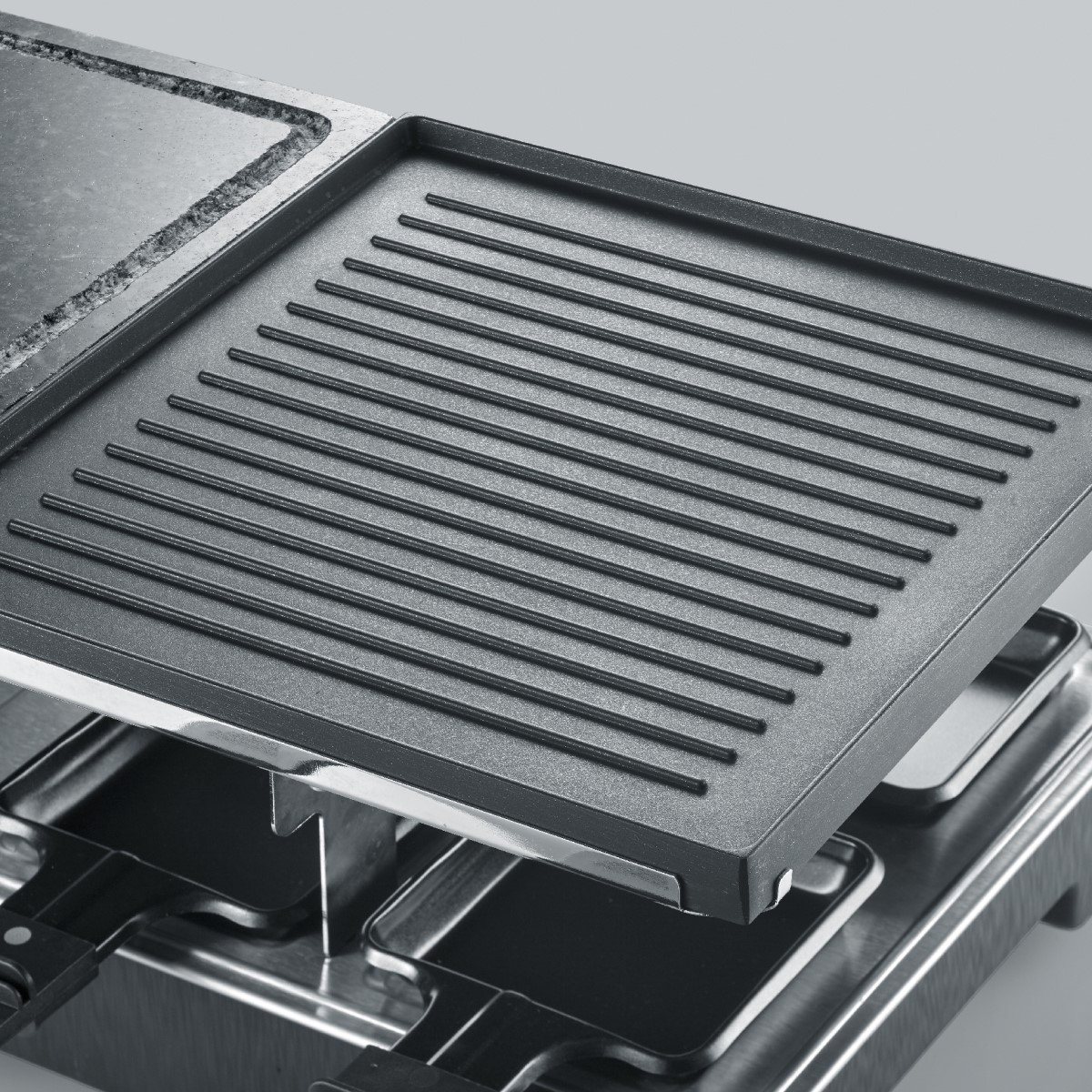 Griglia per raclette con superficie di cottura. Ha un piano grill e spazio per le padelle per il formaggio. I dettagli metallici sono visibili.