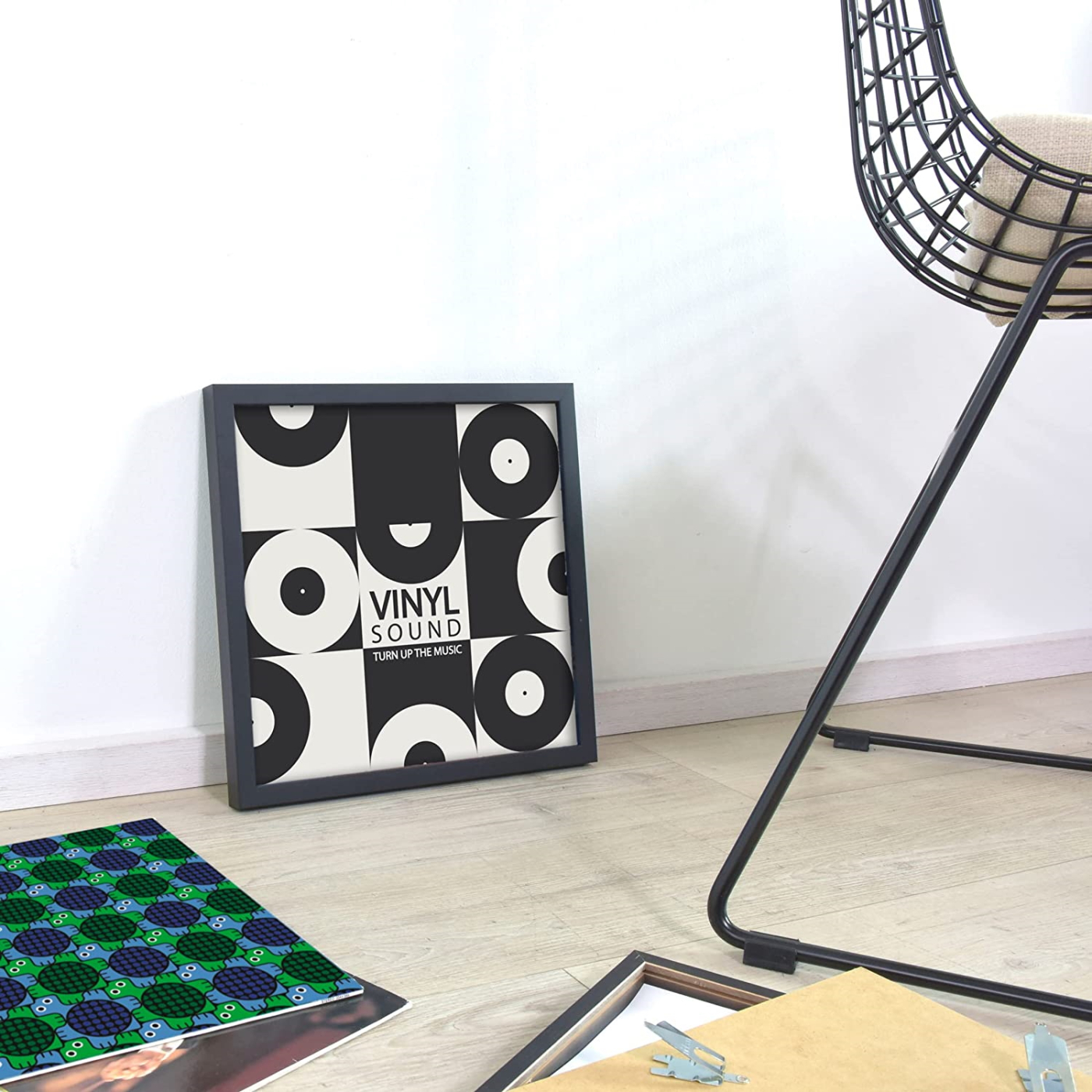 Ein schwarz gerahmtes Poster mit Vinyl-Schallplatten-Design. Ein schwarzer Stuhl, gemustertes Papier und ein Holzboden sind ebenfalls in der Szene zu sehen.