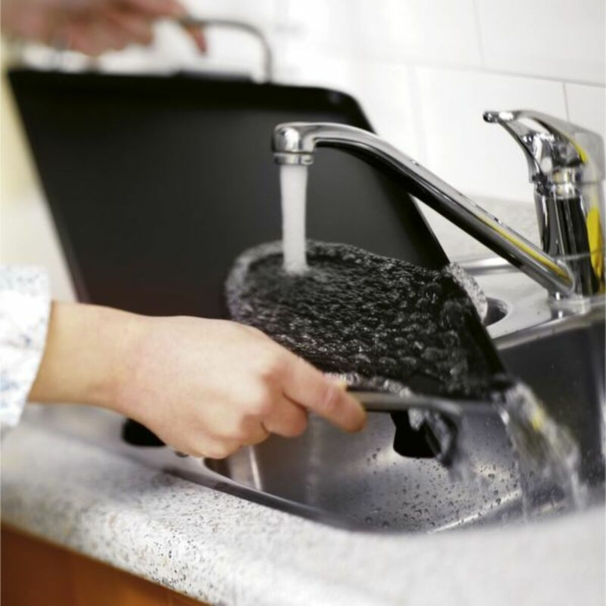 Person wäscht eine schwarze Grillpfanne unter einer Chromarmatur in einem Edelstahlbecken.