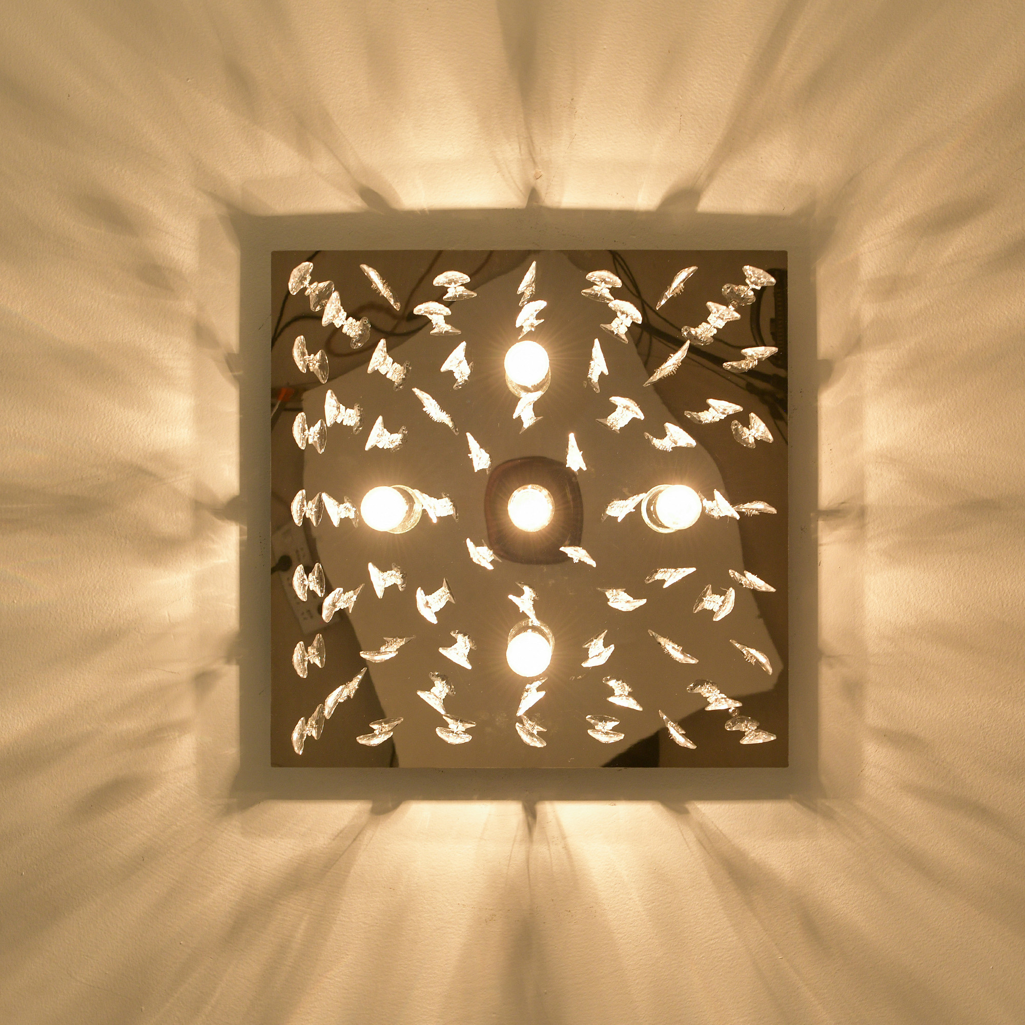 Deckenleuchte mit mehreren Glühbirnen und dekorativem Glas, die Lichtstrahlen auf eine weiße Wand wirft.