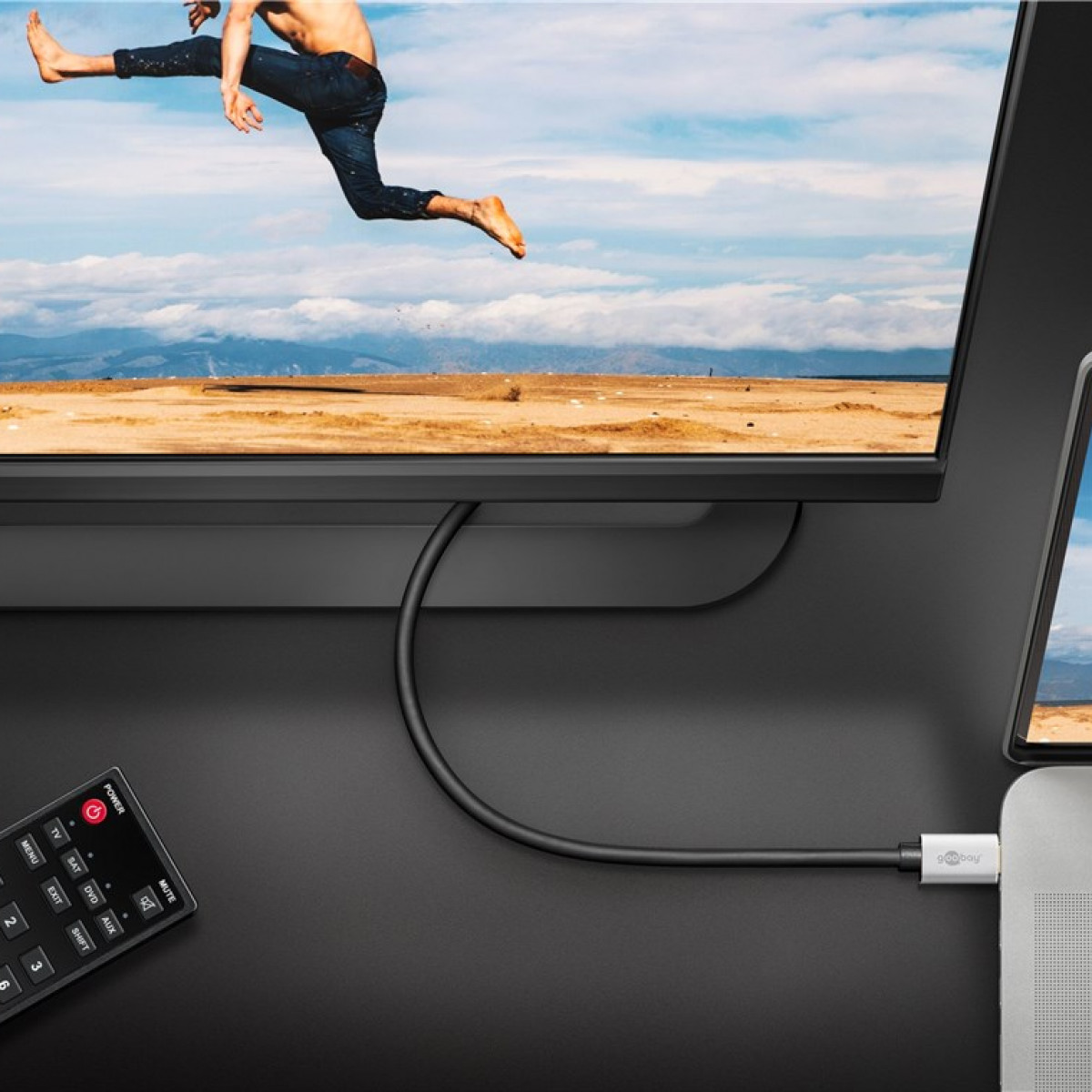 Ein schwarzer Fernseher mit einem Kabel, das mit einem Laptop verbunden ist. Fernbedienung auf schwarzer Oberfläche. Person springt im Hintergrund.