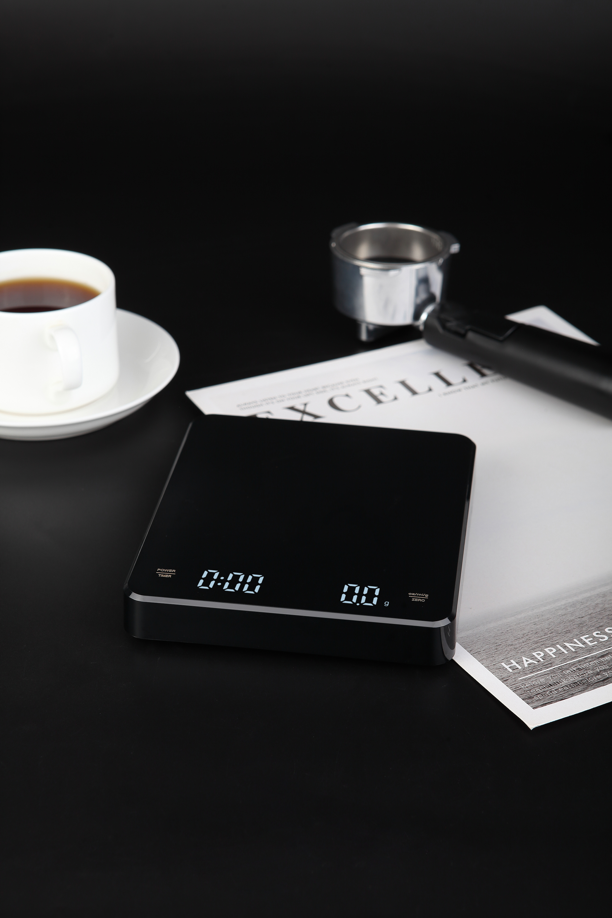 Czarna waga cyfrowa z filiżanką kawy i kilkoma przedmiotami na ciemnej powierzchni.