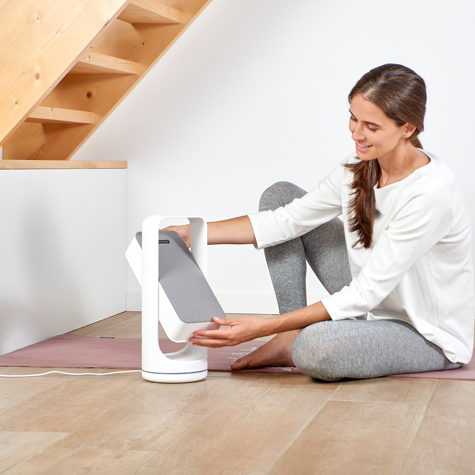 Frau, die ein weißes Lichttherapiegerät verwendet. Sitzt auf einer rosa Matte, in der Nähe einer Treppe. Weißer Hintergrund.