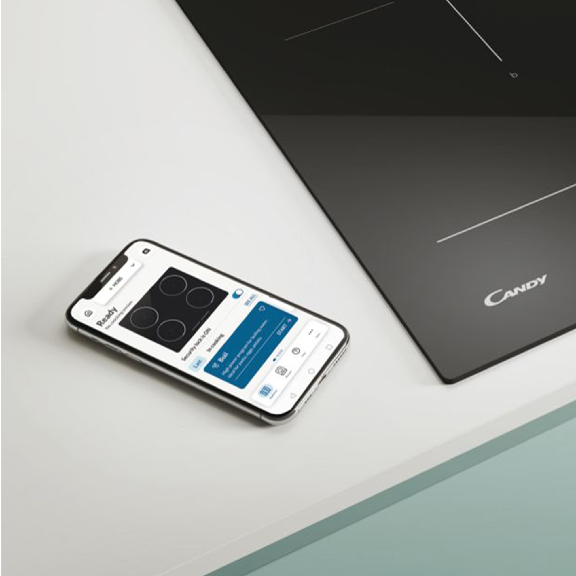 Smartphone mit einer Koch-App neben einem schwarzen Induktionskochfeld. Weiße Oberfläche.